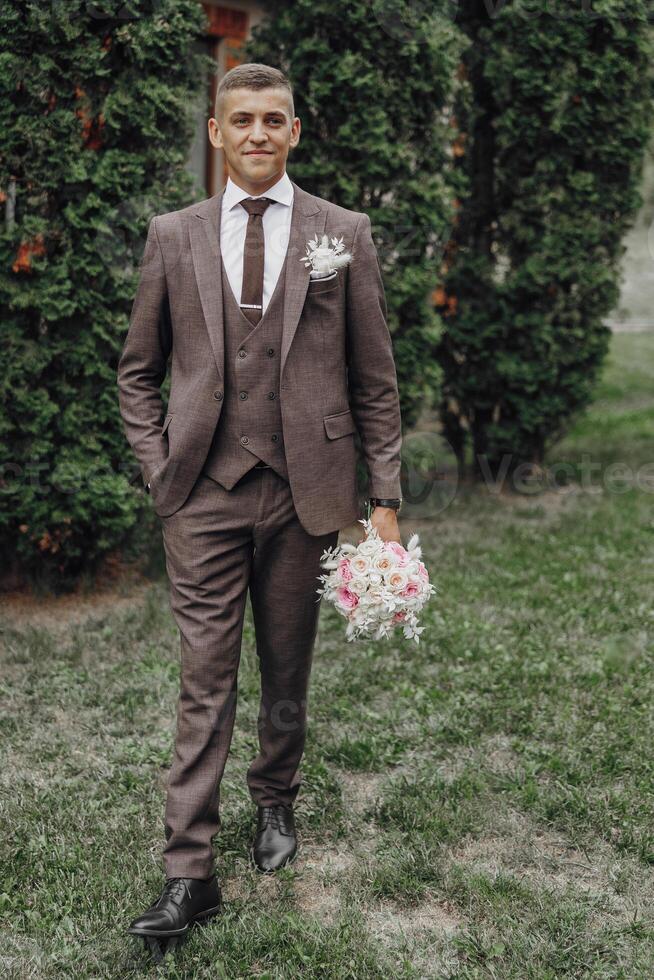 A man in a suit and tie stands in front of a bush holding a bouquet of flowers photo