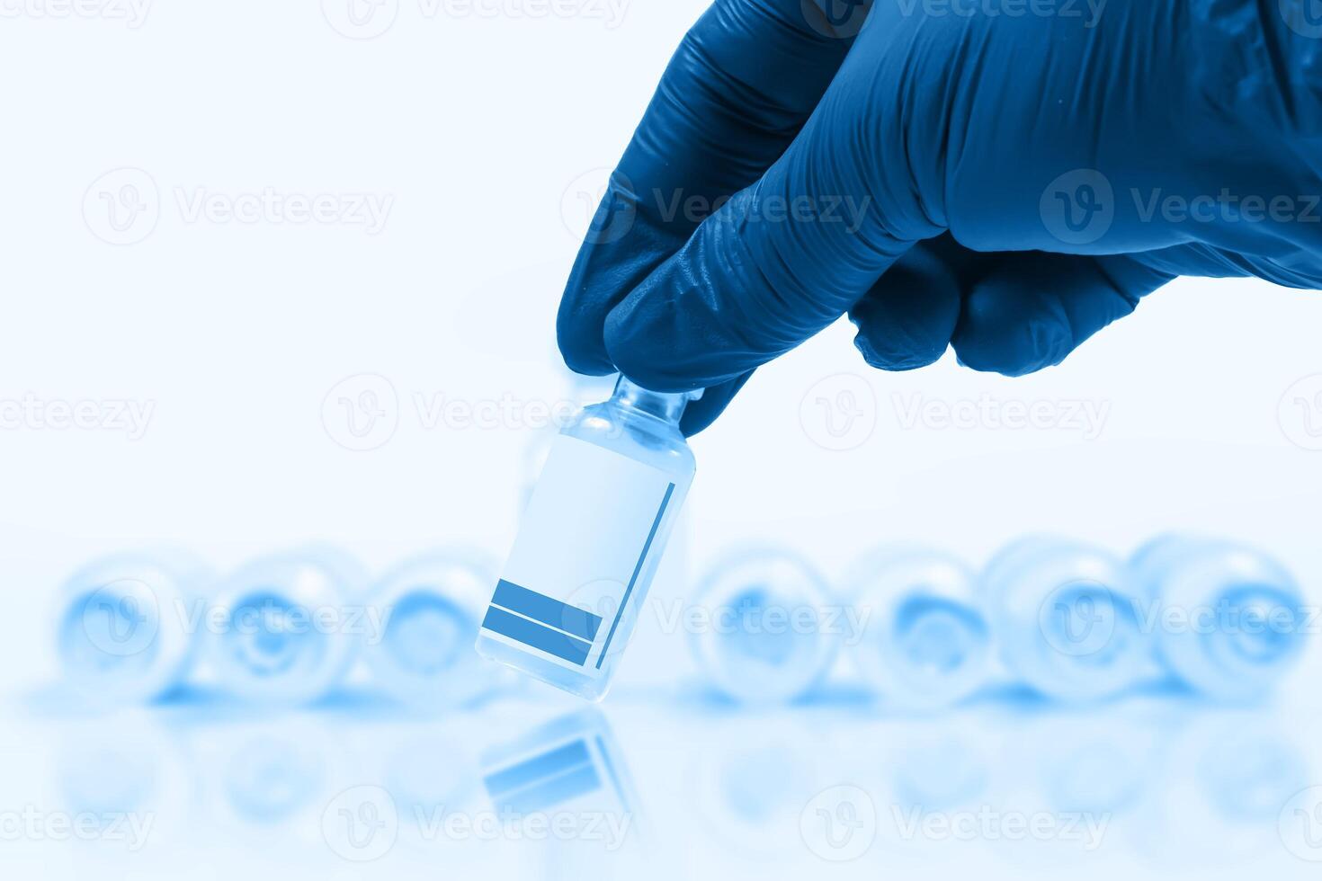 Blue gloved doctor hand holding a vaccine vial in front of a blurred row of vaccine vials on a blue background. photo