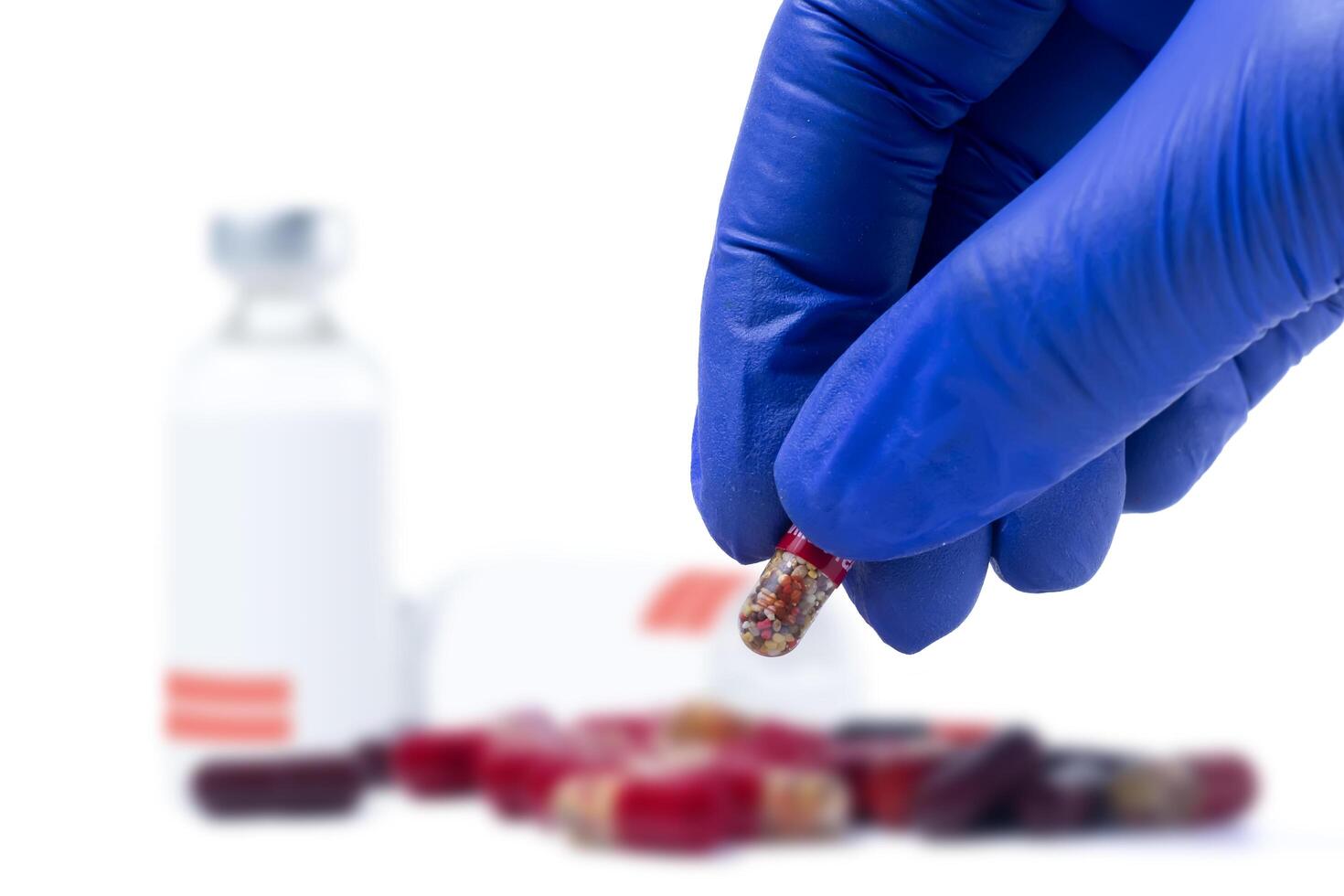 Doctor's hand holding red pill in blue gloves in front of vaccine medical vials and pills with focus outs on white background. Closeup photo