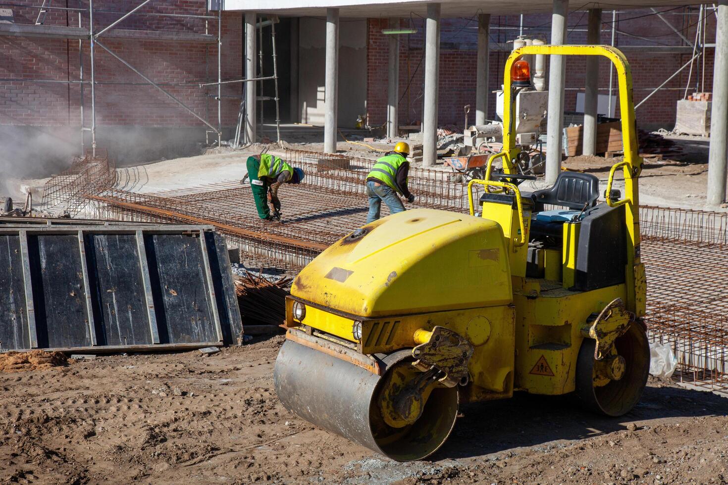 Two construction workers assembling rebar frames at a renovation site, with a compactor in the foreground. Safety helmets and reflective vests emphasize the professional work environment. photo