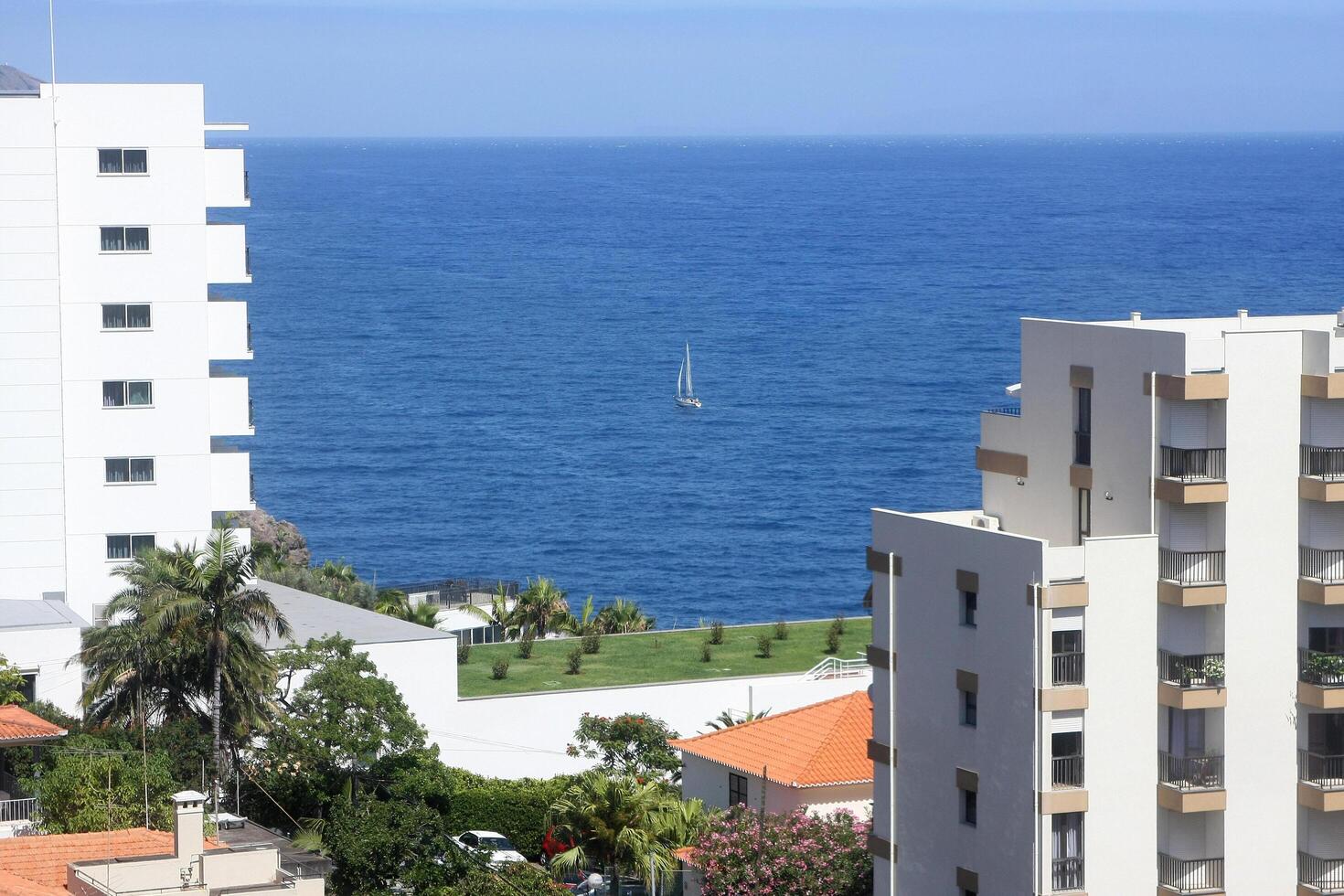 A small sailing boat glides gracefully between two buildings in Funchal, Madeira Island, framed by a clear sky and shimmering waters, capturing a serene maritime moment. photo