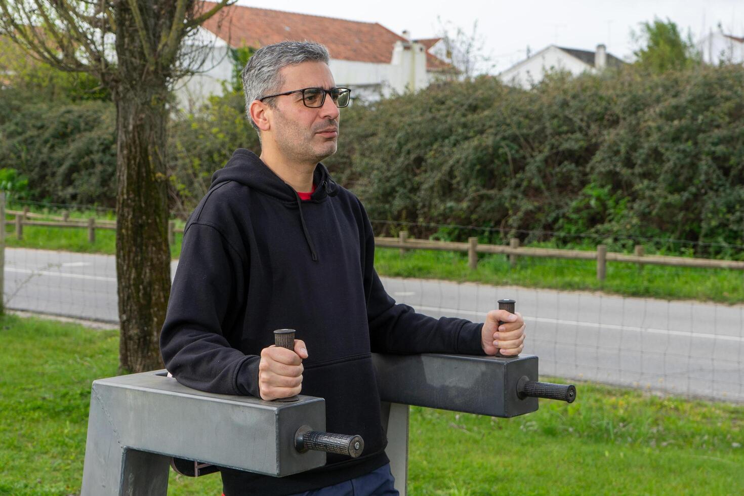 A gray-haired man performs crunches on an exercise machine in an urban park, showcasing fitness and an active lifestyle amidst a green, city environment. photo