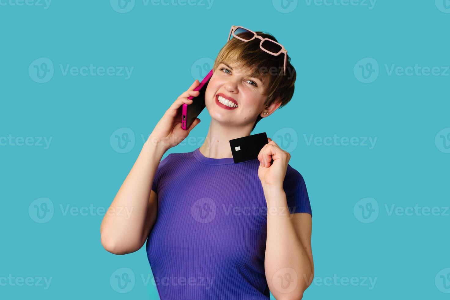 The concept of online shopping. A Caucasian young woman holds a credit card, talks on a smartphone, makes a purchase, orders delivery, standing on a blue background. photo