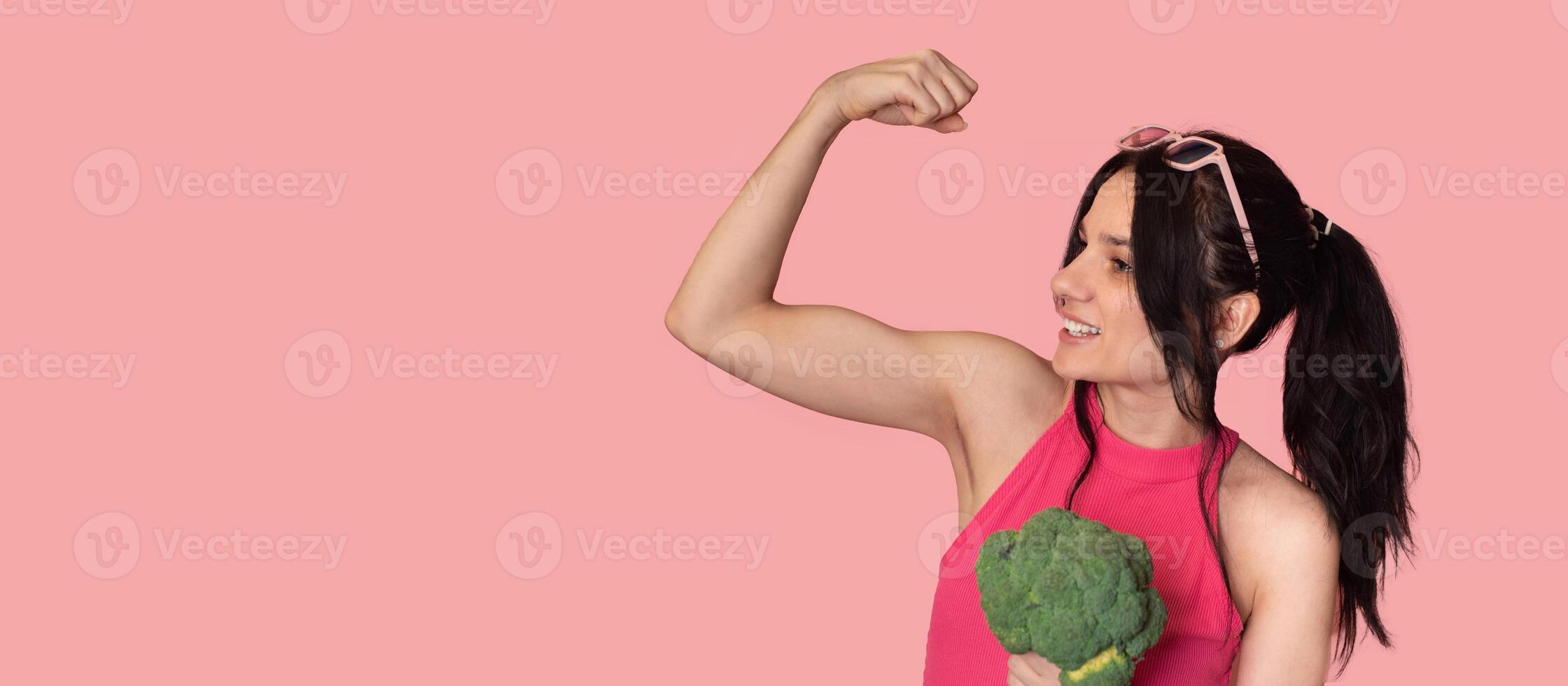 A smiling white girl with fresh broccoli in one hand and showing off her biceps in the other. Panoramic banner, free space. photo