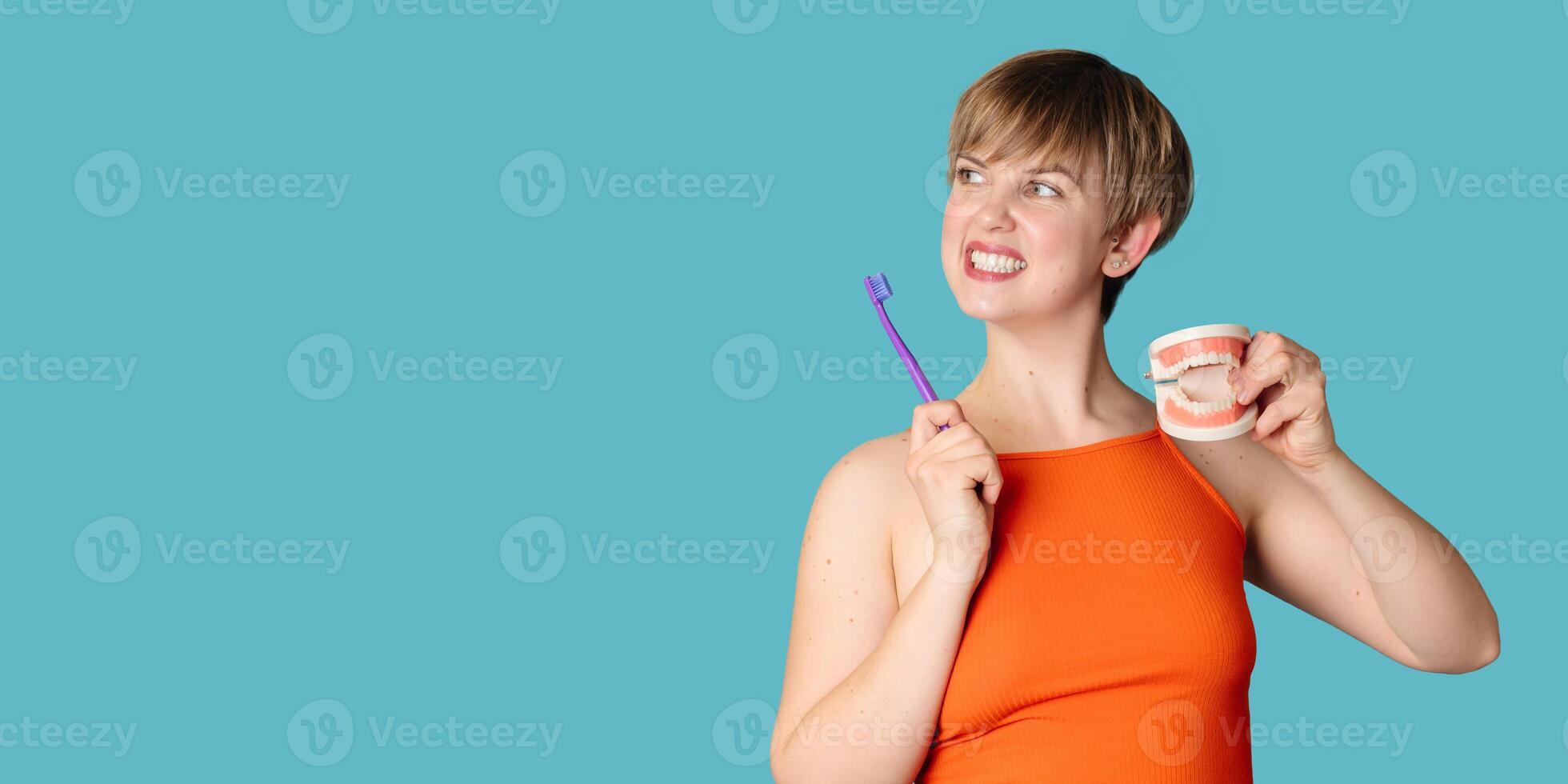 A young white woman with an amazing smile, a toothbrush and a jaw model in her hands on a blue background. A place for your text. photo