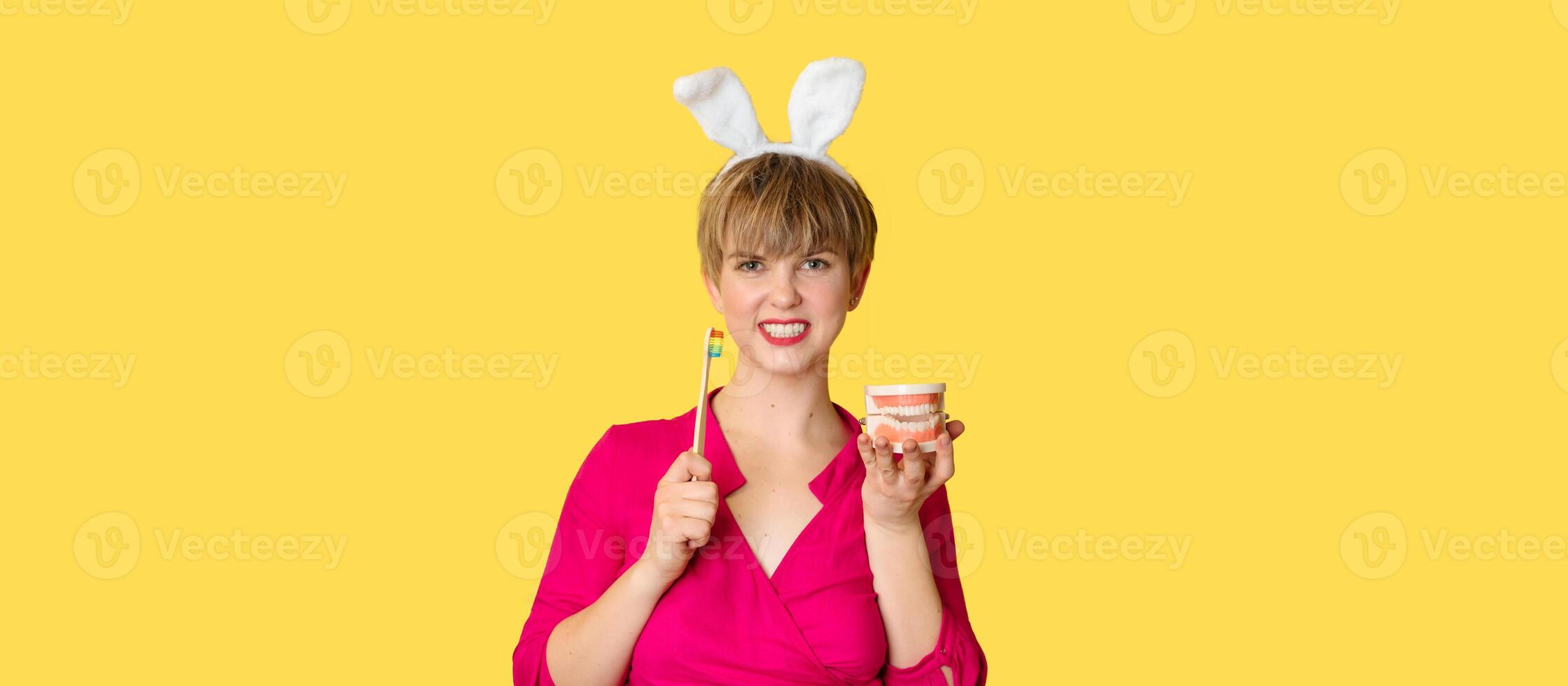 A young woman in bunny ears with a toothbrush and a jaw model in her hands on a yellow background. A place for your text. photo