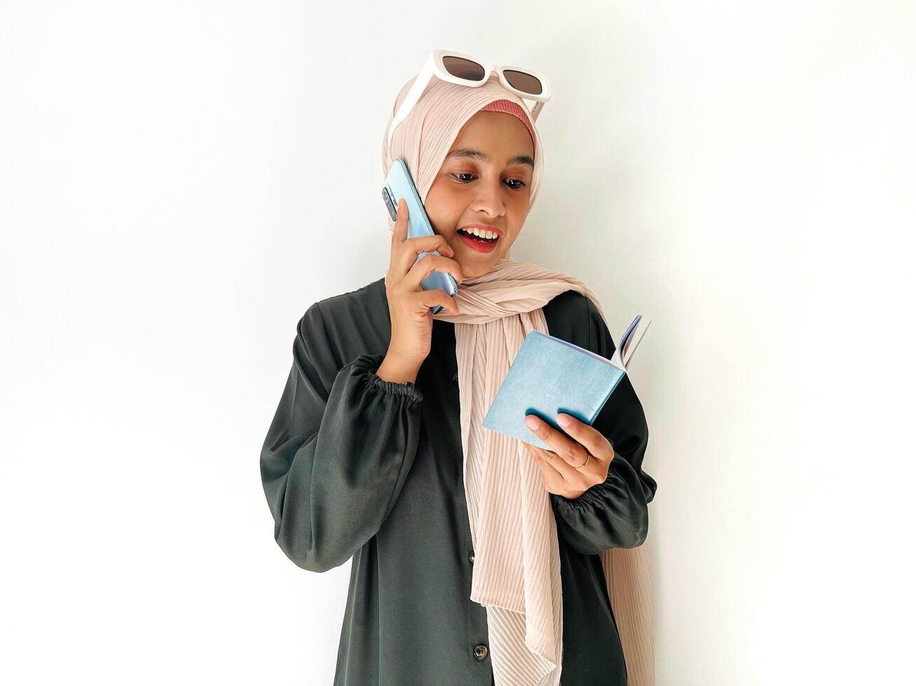 A standing and smiling woman wear sun glasses on her head holding her cell phone and holding Indonesian passport while looking at an Indonesian passport with an open page isolated on white background photo