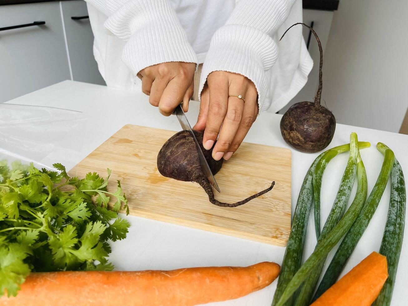 Cooking concept. Unrecognizable woman in white clothes making healthy dinner, cropped of lady preparing delicious meal at home, cutting beetroot vegetables on chopping board, kitchen interior photo