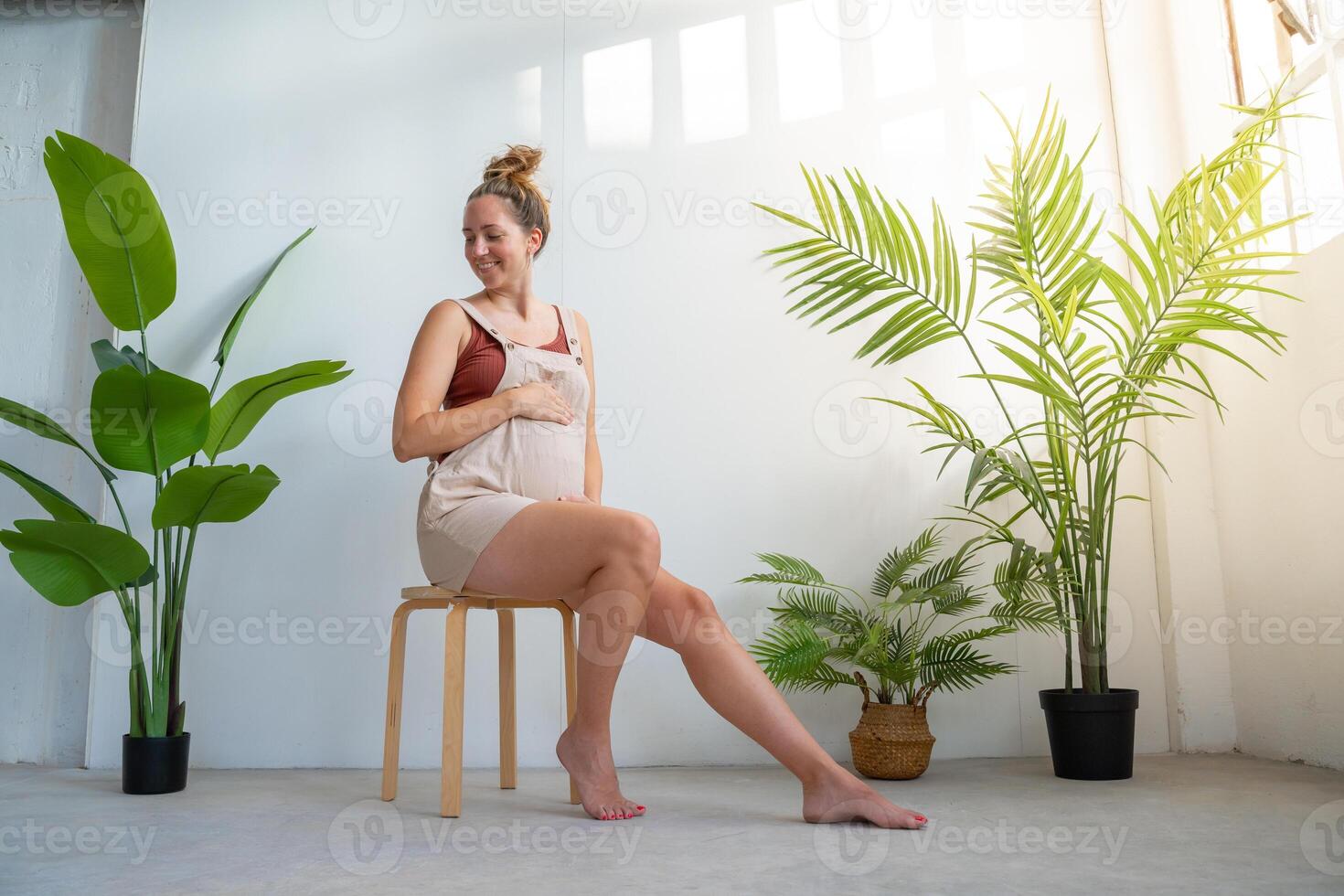 Happy pregnant women touching her belly relaxed on a chair between indoor plants. photo