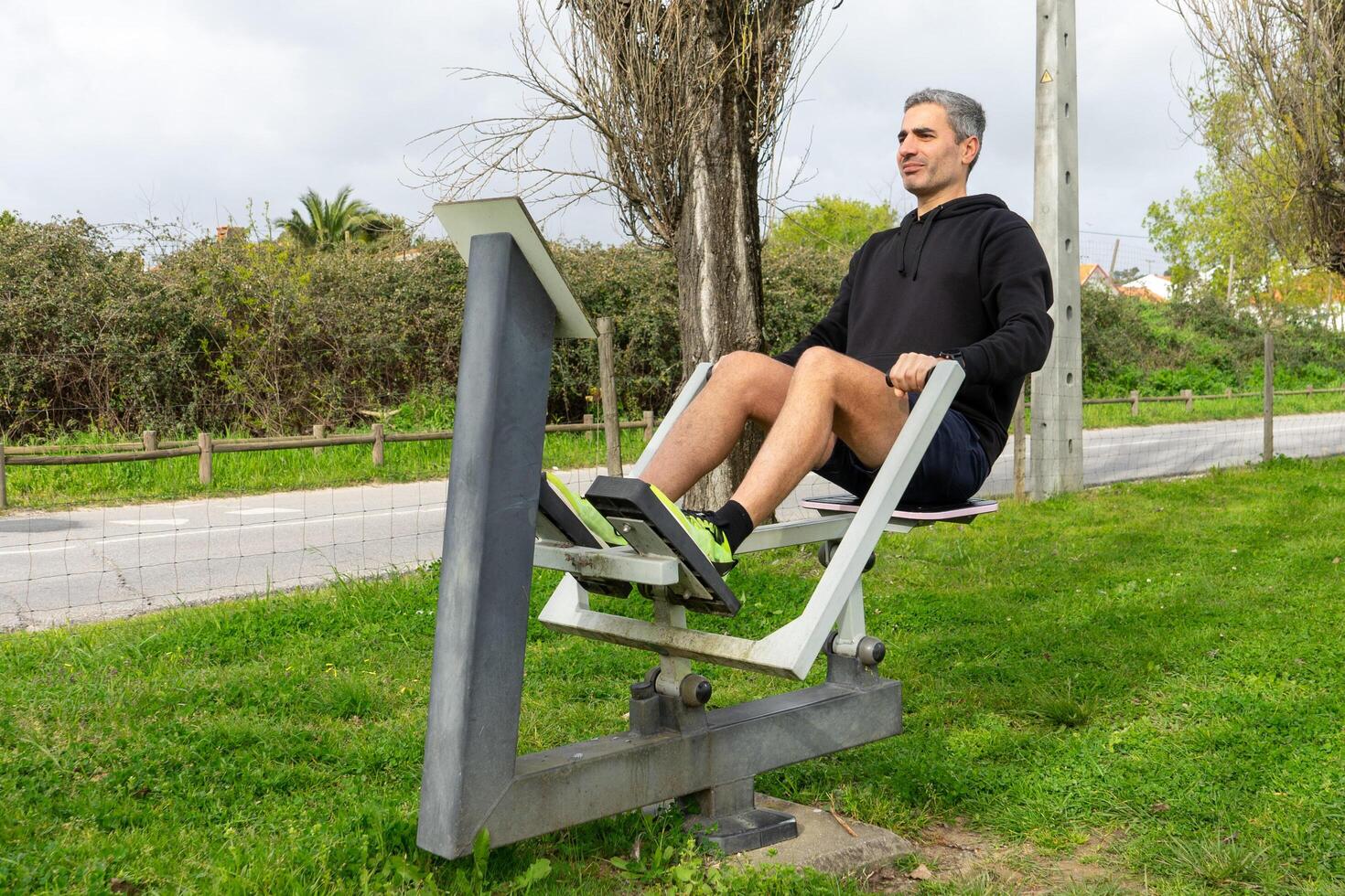 A grey-haired man working out on a rowing machine in an outdoor fitness circuit in an urban park, surrounded by greenery. photo
