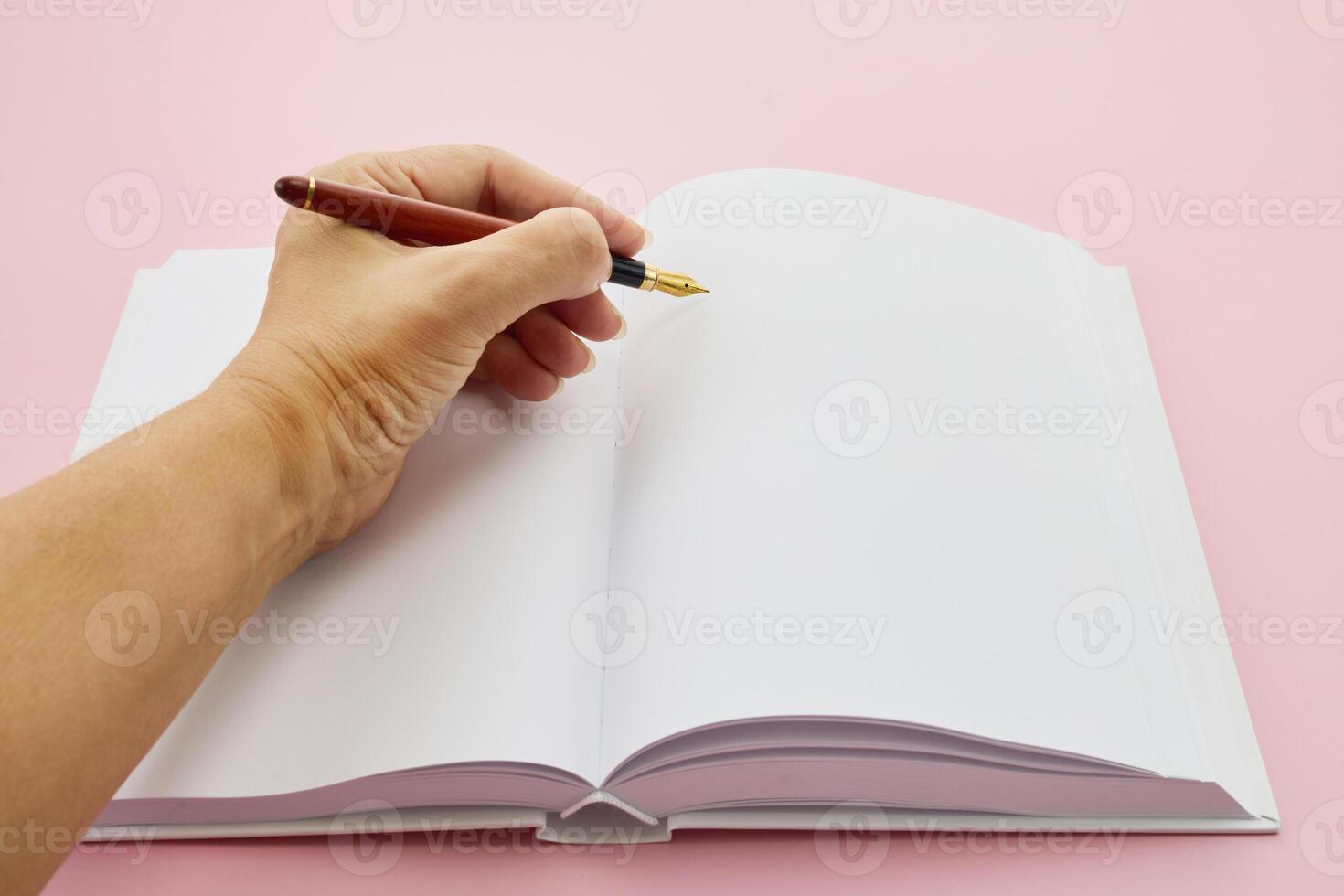 International Left-handers Day. Left hand holding a pen, writing in an open blank journal on a light pink background. photo