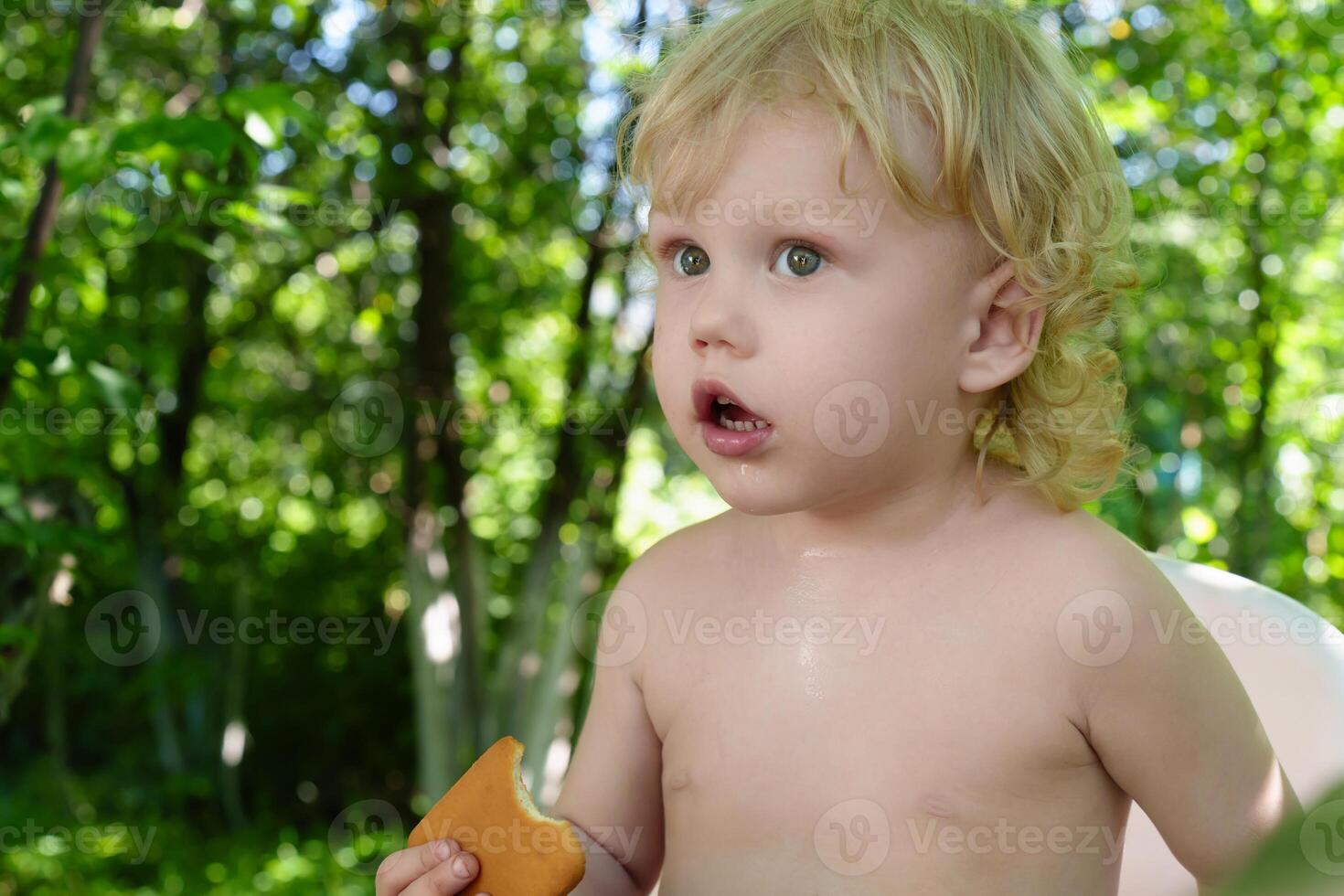 Curly blond toddler happily nibbling on a freshly baked cookie while sitting in aagainst a backdrop of garden trees on a summer day photo