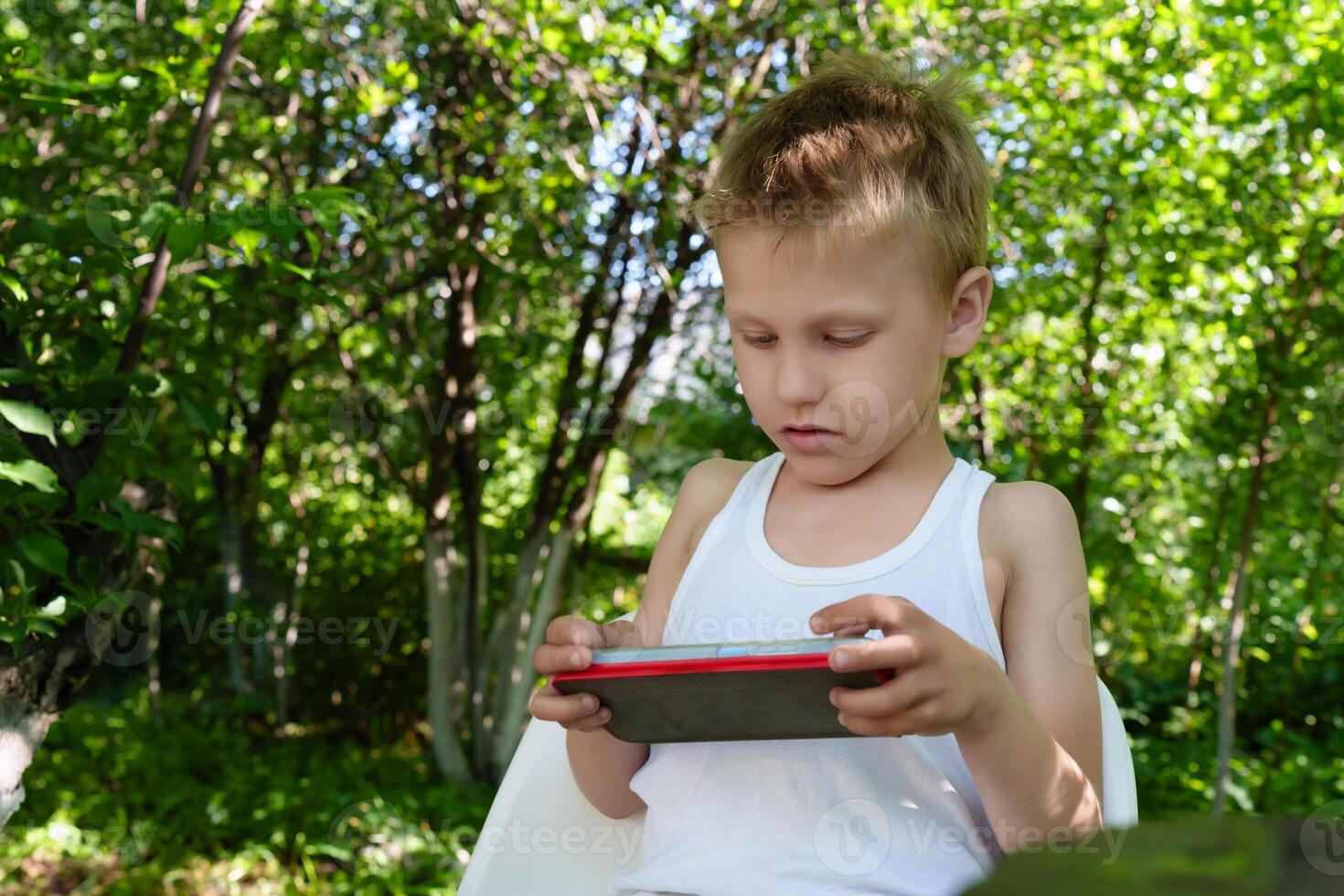 Young boy sits amidst lush greenery, his attention fully absorbed by the interactive games displayed on the screen of his mobile device photo