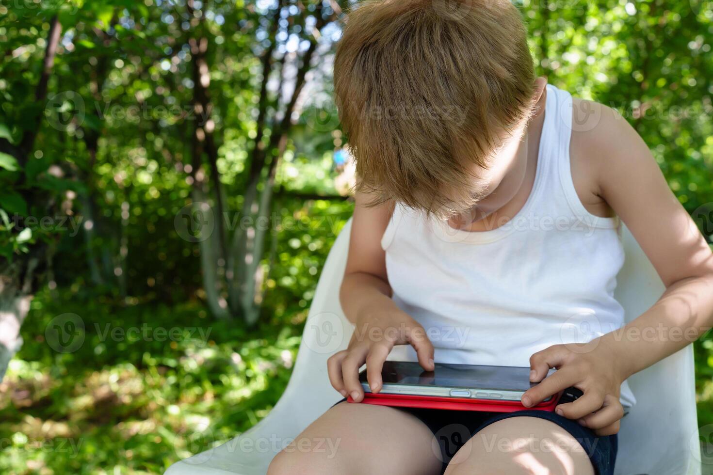 Solitary child with fair locks sits hunched over, absorbed in the digital world of his cell phone photo