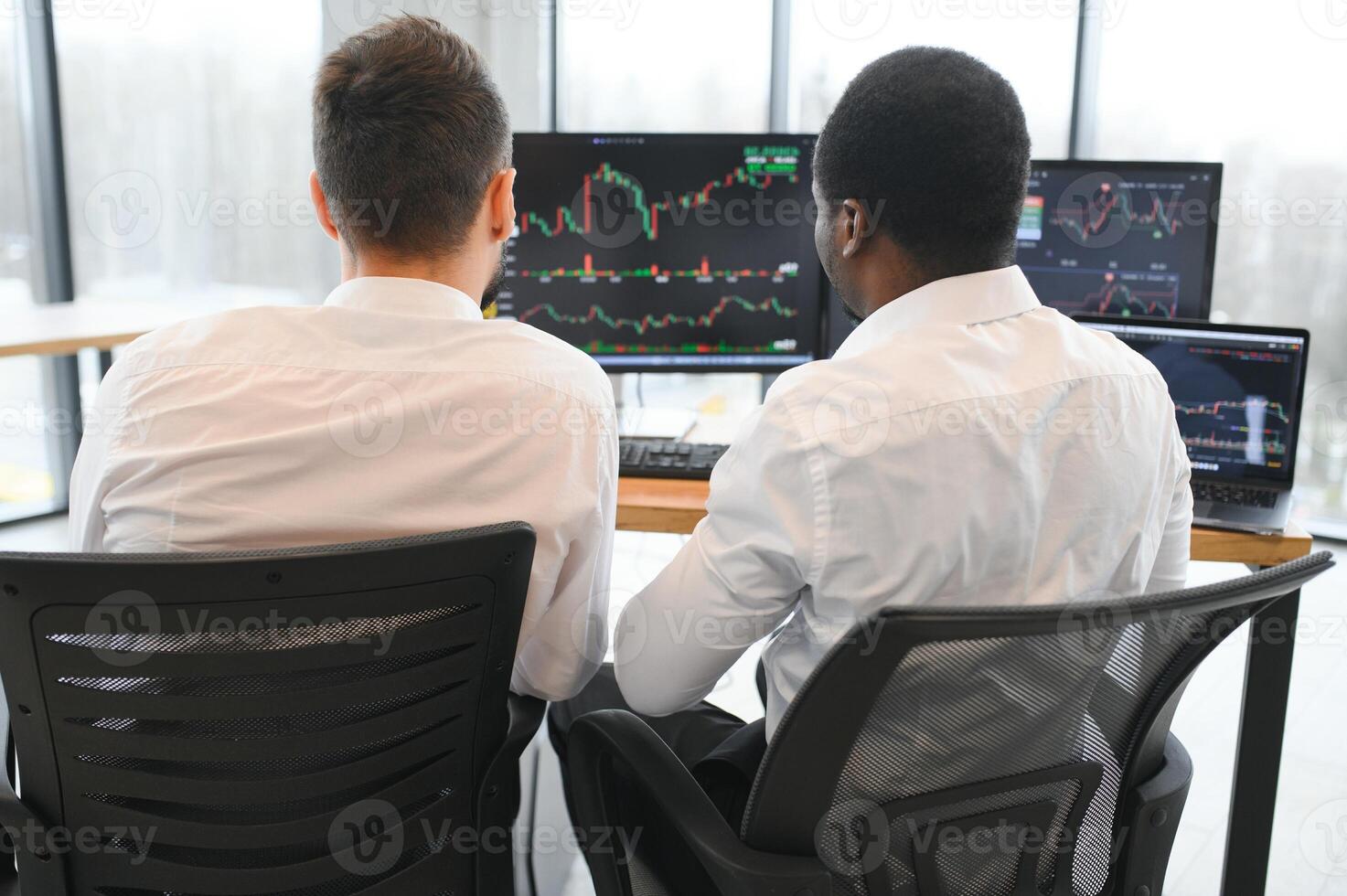 Two diverse colleagues traders talking to each other, sitting in the office in front of multiple computer screens. Stock trading, people, business concept photo