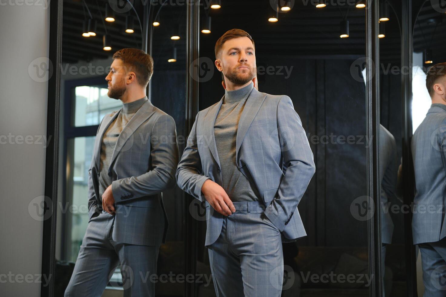 Modern young handsome businessman dressed in classical suit adjusting a tie in front of the mirror while standing in the suit shop photo
