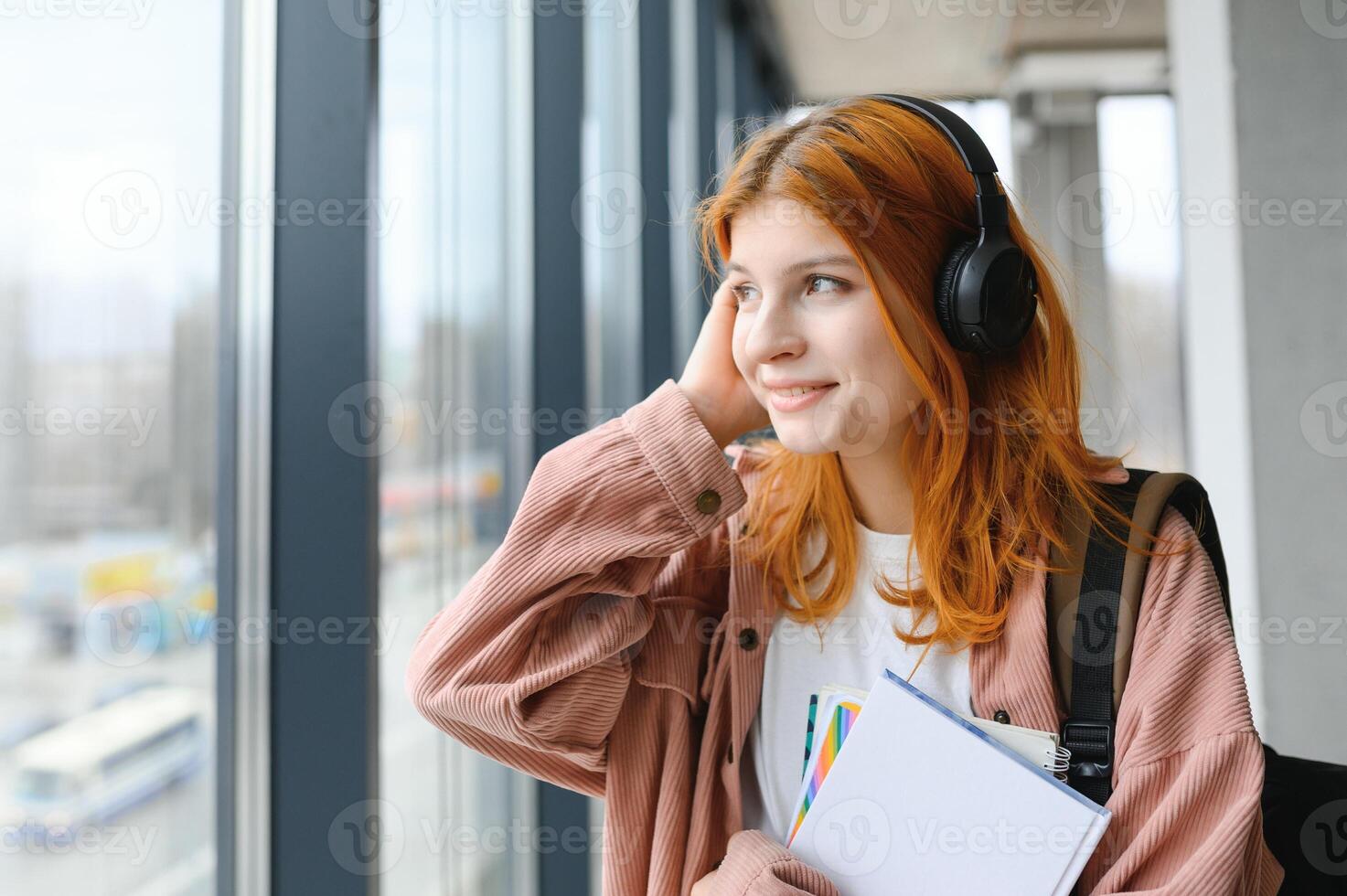 Red-haired student girl stands by the window with books and a backpack photo