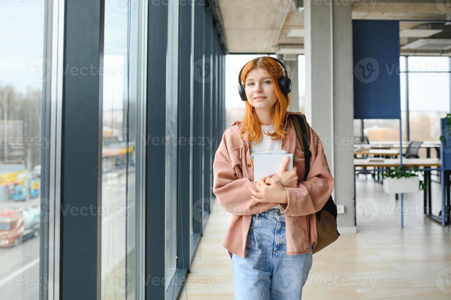 Red-haired student girl stands by the window with books and a backpack photo