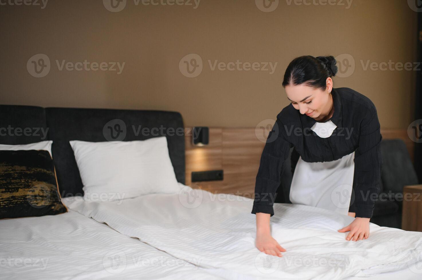 Young pretty housekeeper in uniform changing bedclothes in hotel room while standing by double bed photo