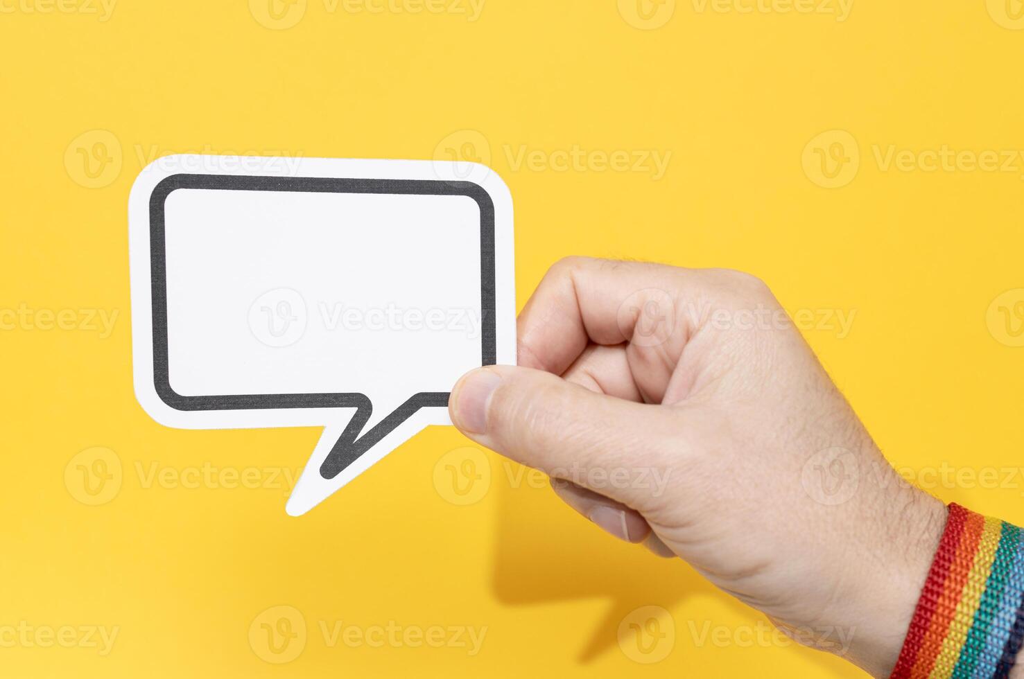 Hand wearing pride flag bracelet holding blank speech bubble isolated on yellow background. Template Mockup photo