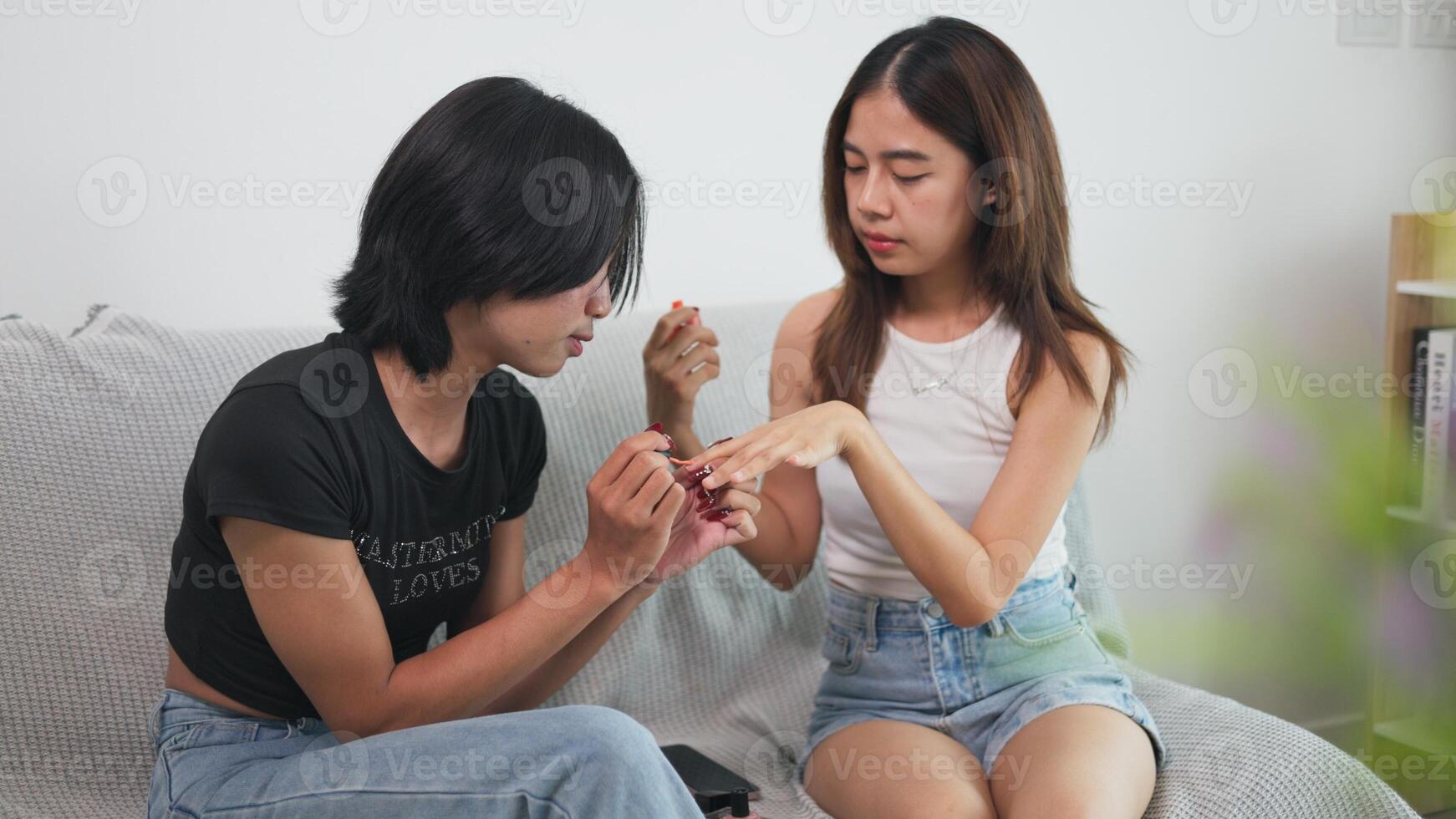 Girl and LGBTQ friend sitting together on sofa, one painting the other's nails in their preferred style. photo