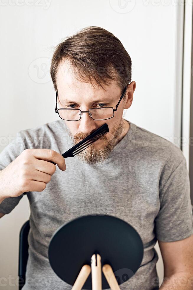 An attractive middle-aged man straightens his beard in front of a mirror. Natural look, light blond hair and freckles, beard with gray hair. Soft focus photo