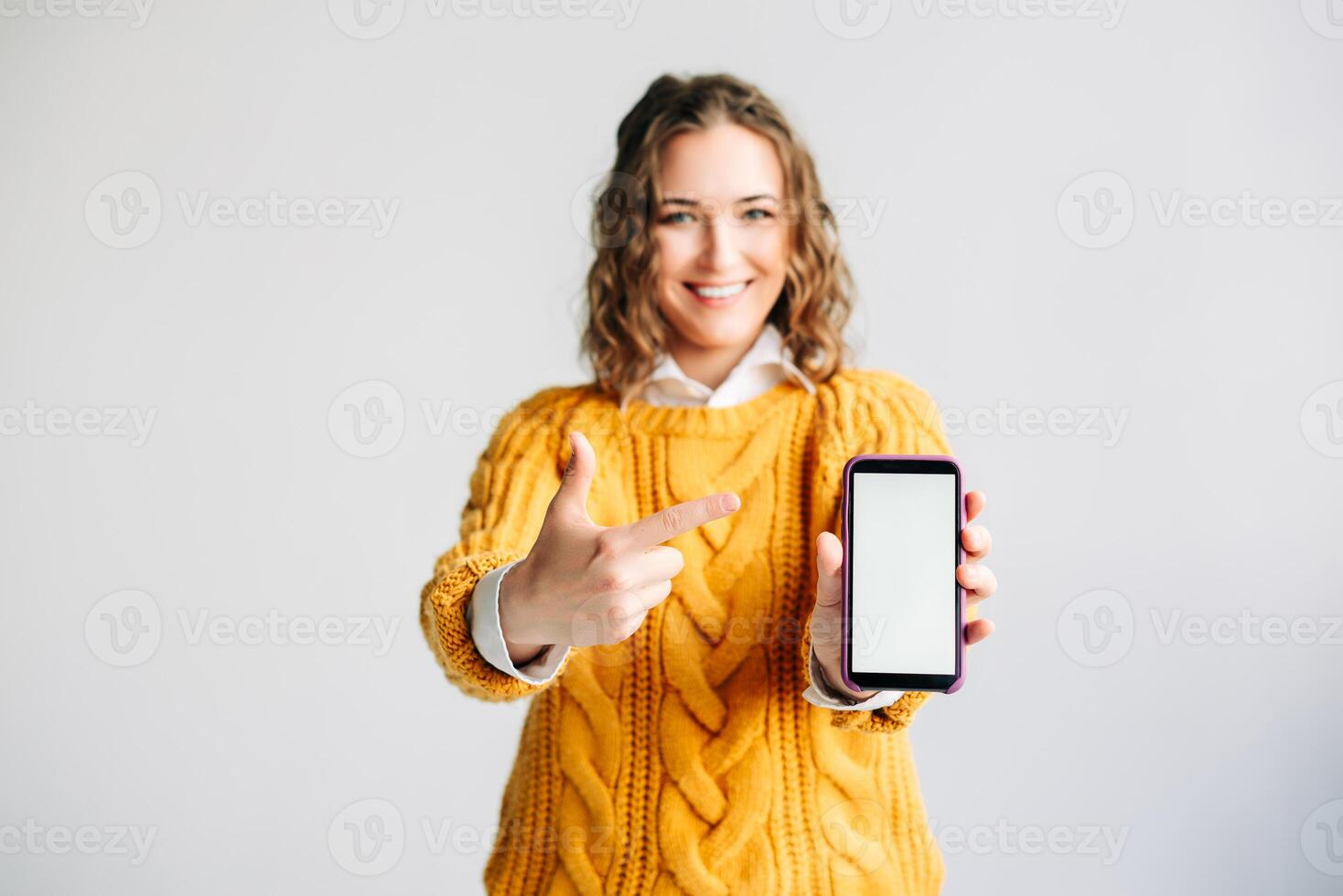 Smiling Woman Pointing at Mobile Phone Screen. Happy woman pointing at mobile phone screen, showcasing a new app. Holding a smartphone with a blank display, isolated on white background. photo
