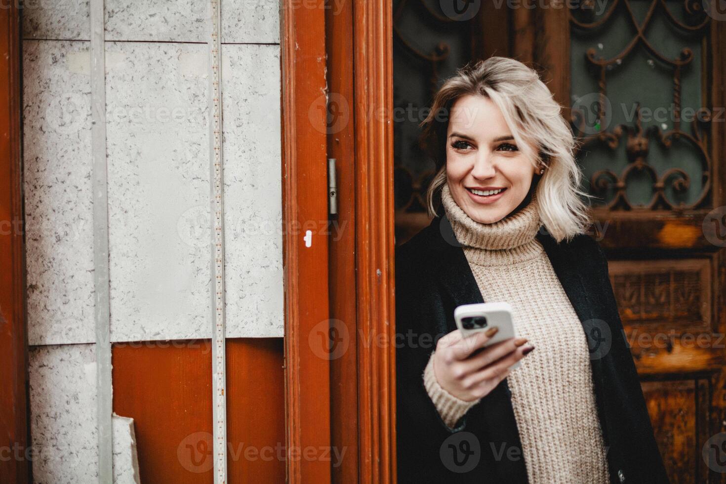 Smiling Woman in Black Coat with Smartphone. Cheerful woman with a black coat and white sweater, holding a smartphone. Ideal for concepts related to digital communication, professional networking photo