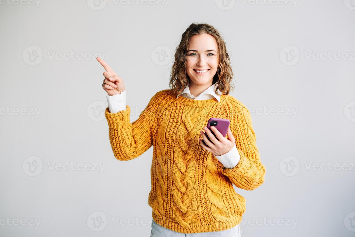 Smiling Young Woman Using Phone, Pointing Aside. A happy young woman model smiles while using her phone, pointing aside against a white background. She presents a new app, advertises mobile web photo