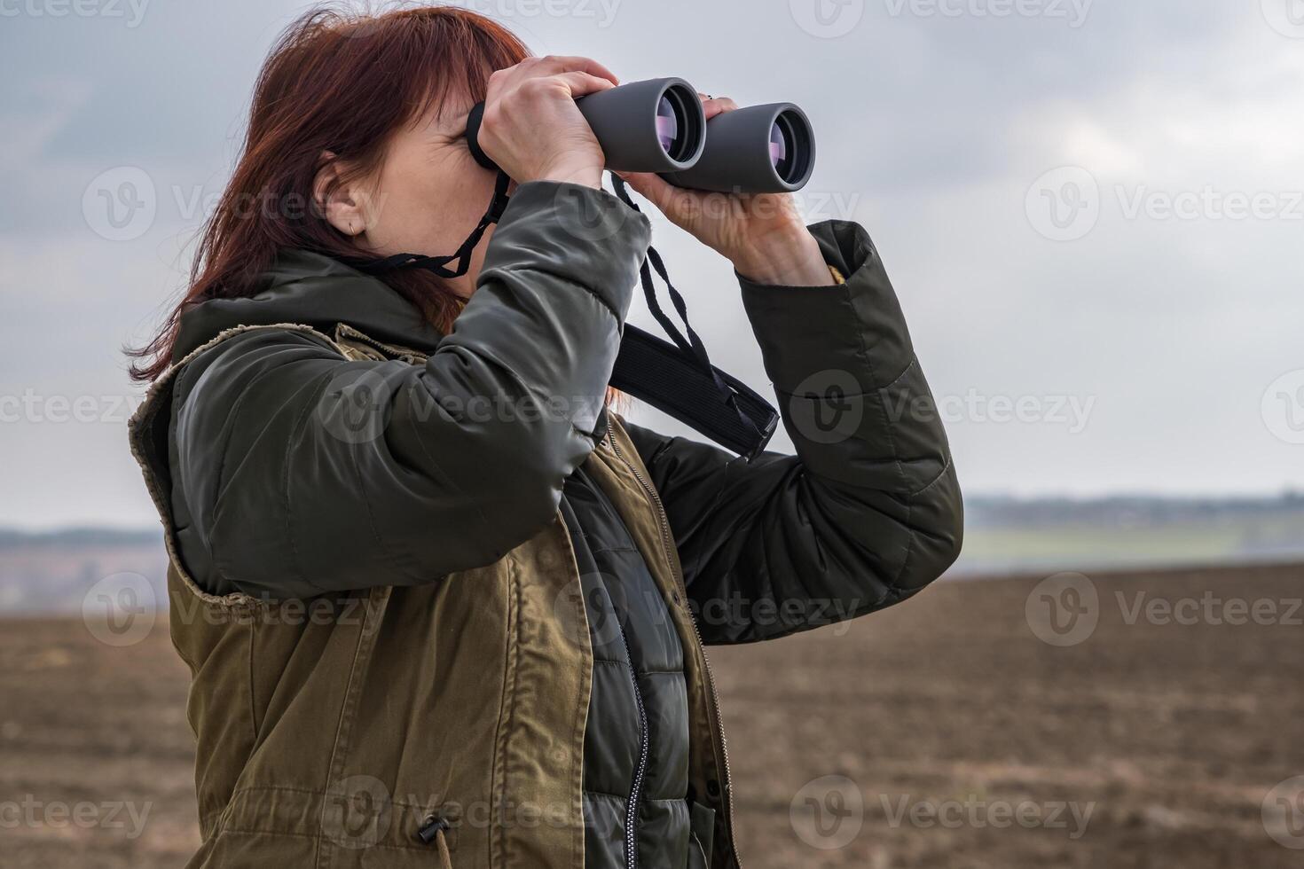 female ornithologist birdman watches birds with binoculars on dirt gravel road against a background of a stormy sky photo
