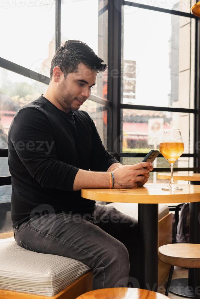 Young Latino man using cell phone in a bar, drinking craft beer at a table. photo
