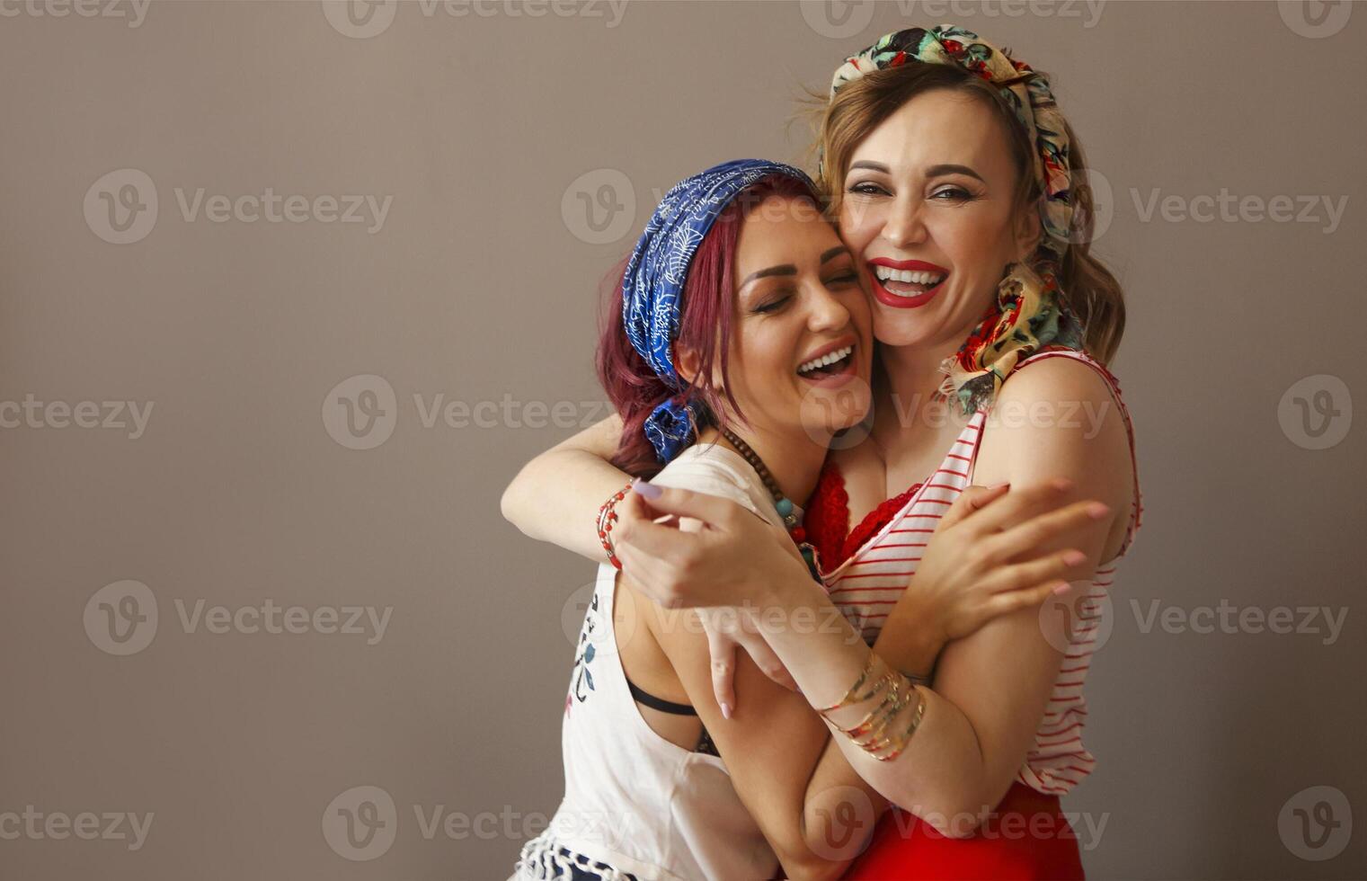 Portrait of two beautiful women in bright clothing and bow in head with bright lips hugging in studio on brown background photo