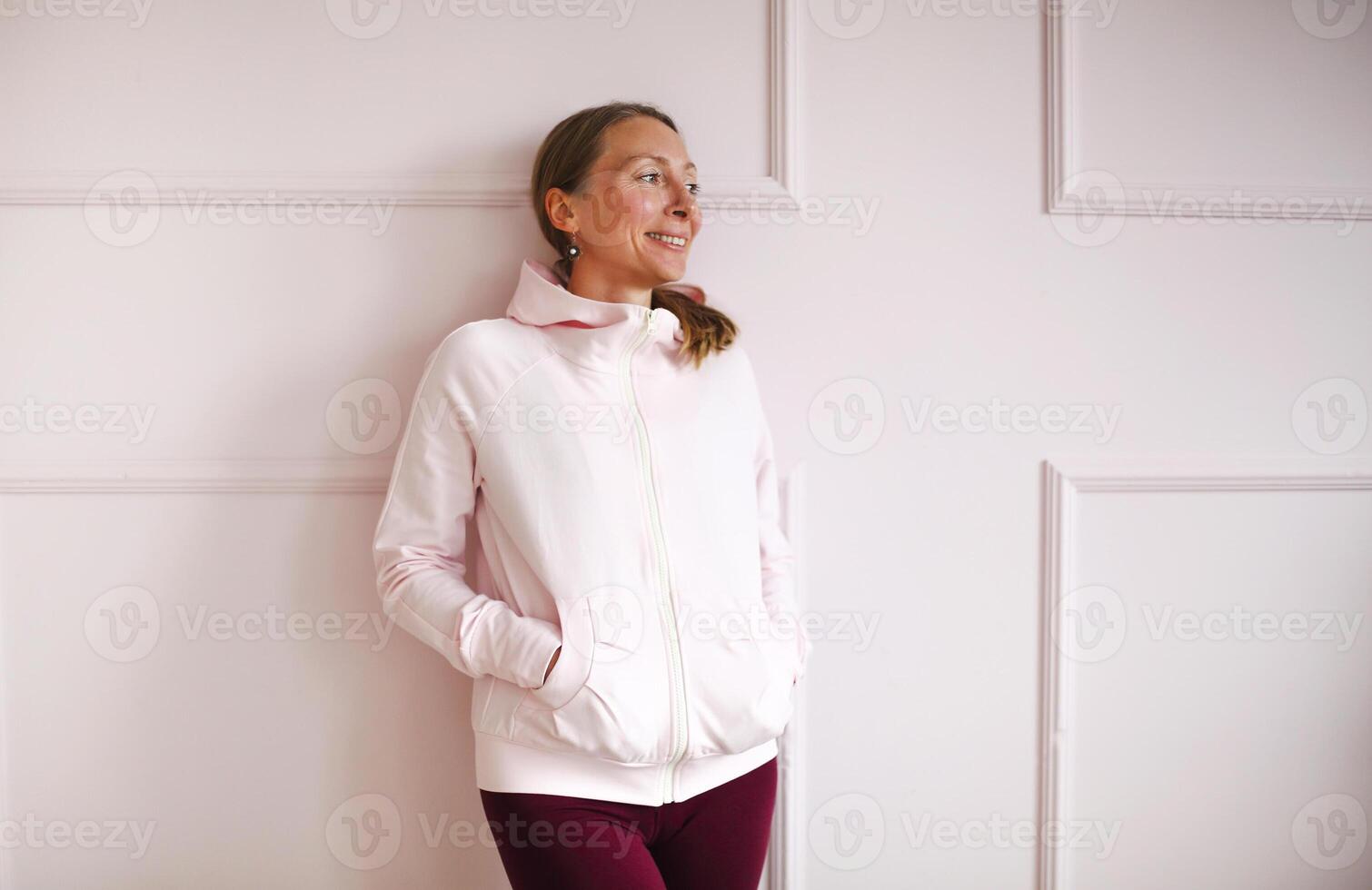 Happy joyful 40s female in sportive pink hoody standing with smile against white wall in gym photo