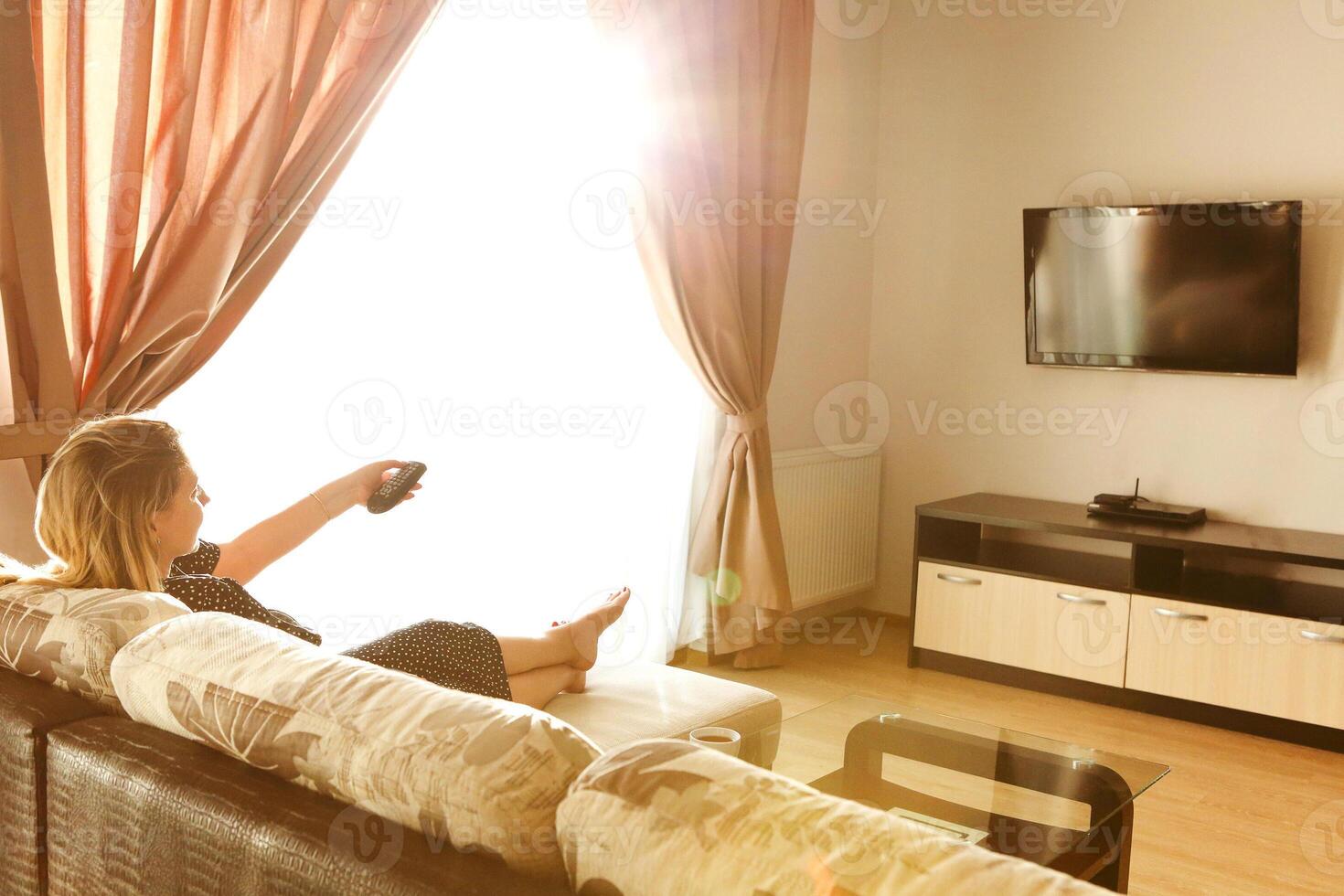 Back view of young woman watching tv while resting and sitting on sofa in living room photo