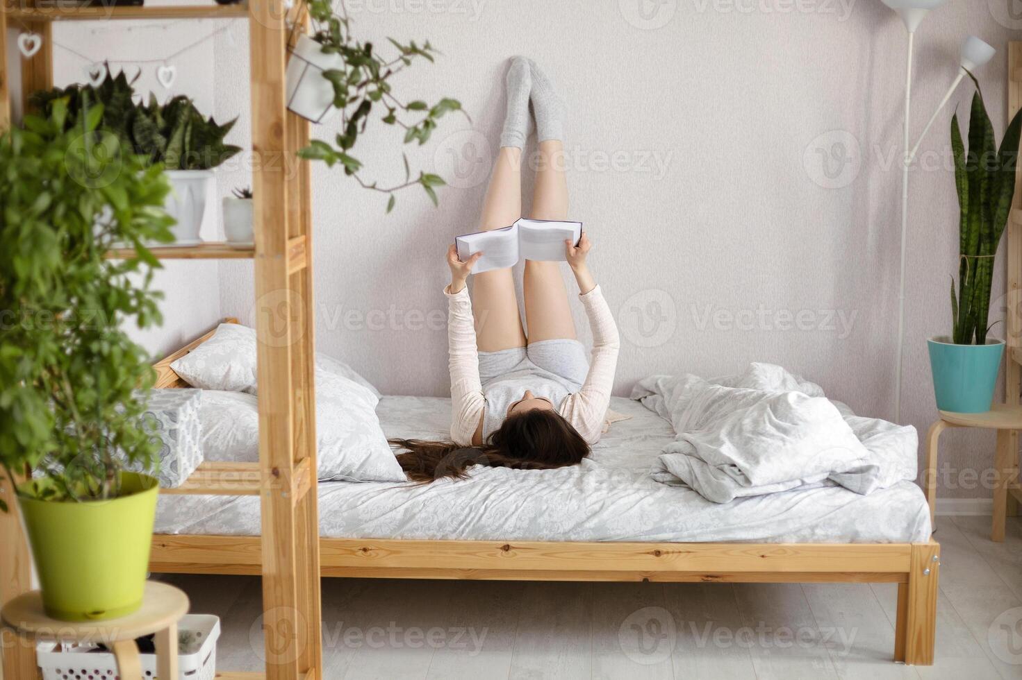 Young brunette girl lying down on bed with legs raised up on wall while holding and reading book photo