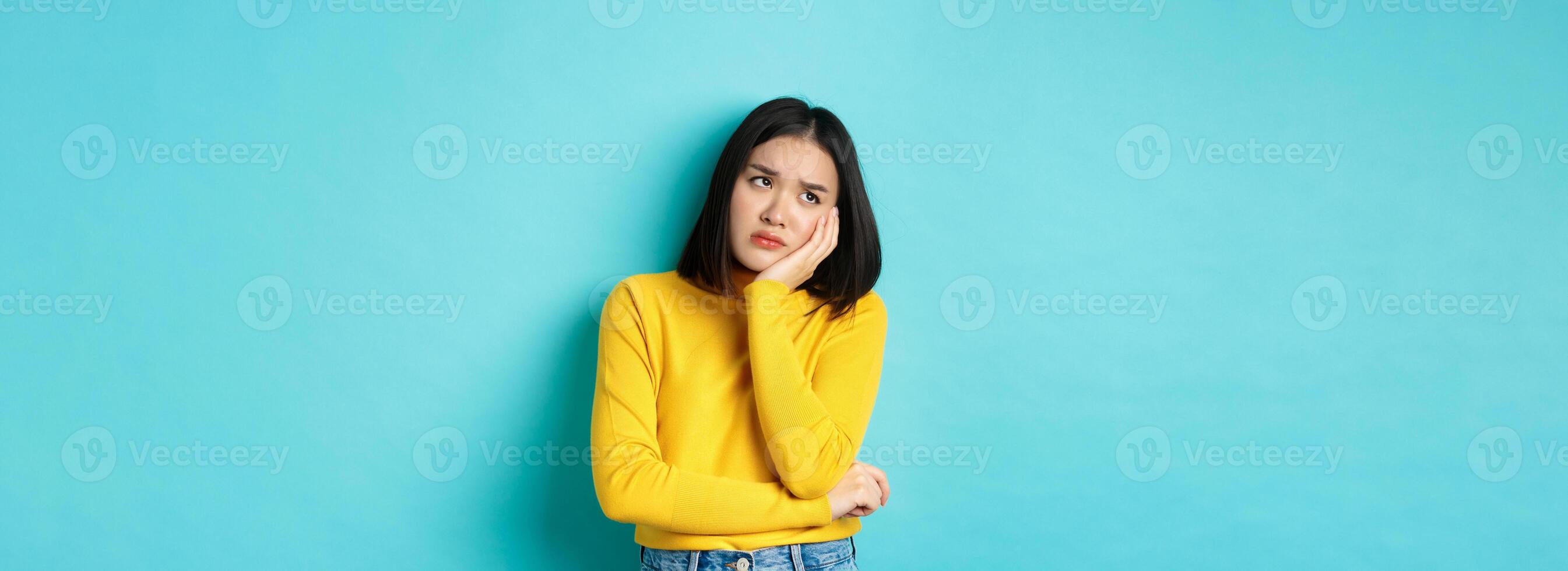 Sad and bored asian teenage girl leaning on palm, looking at upper left corner with lonely and upset expression, standing over blue background photo