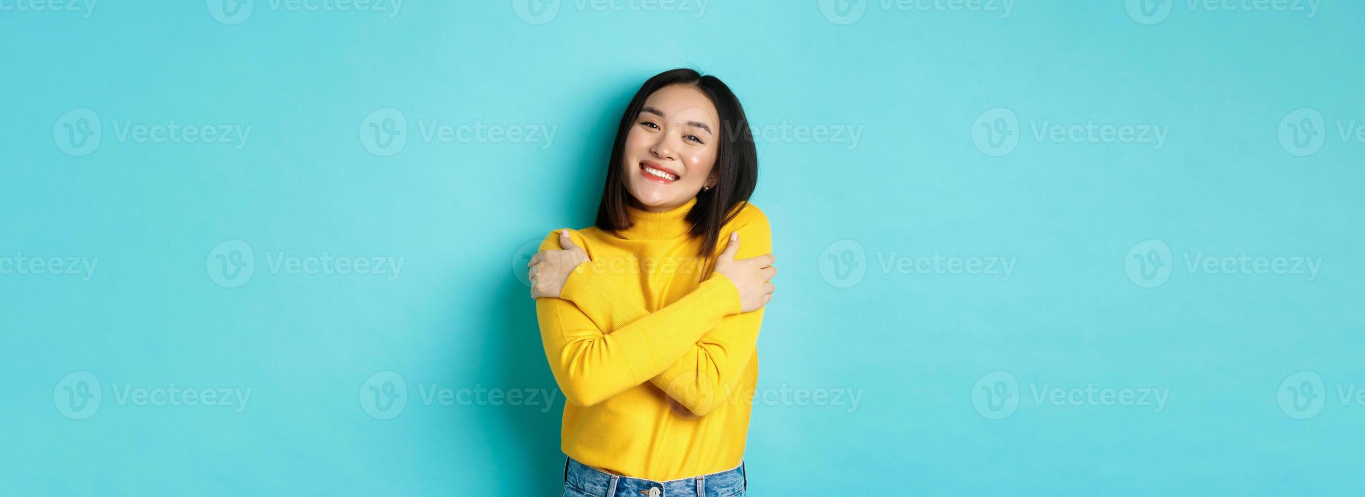 Cheerful beautiful girl embracing own body, smiling and cuddling herself, standing carefree in yellow pullover against blue background photo