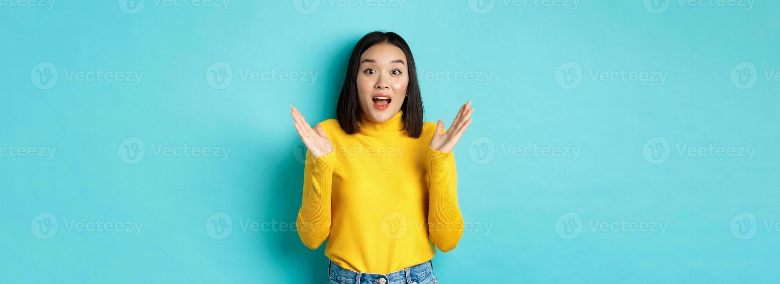 Image of happy asian woman clap hands and looking amazed, standing in yellow pullover over blue background photo