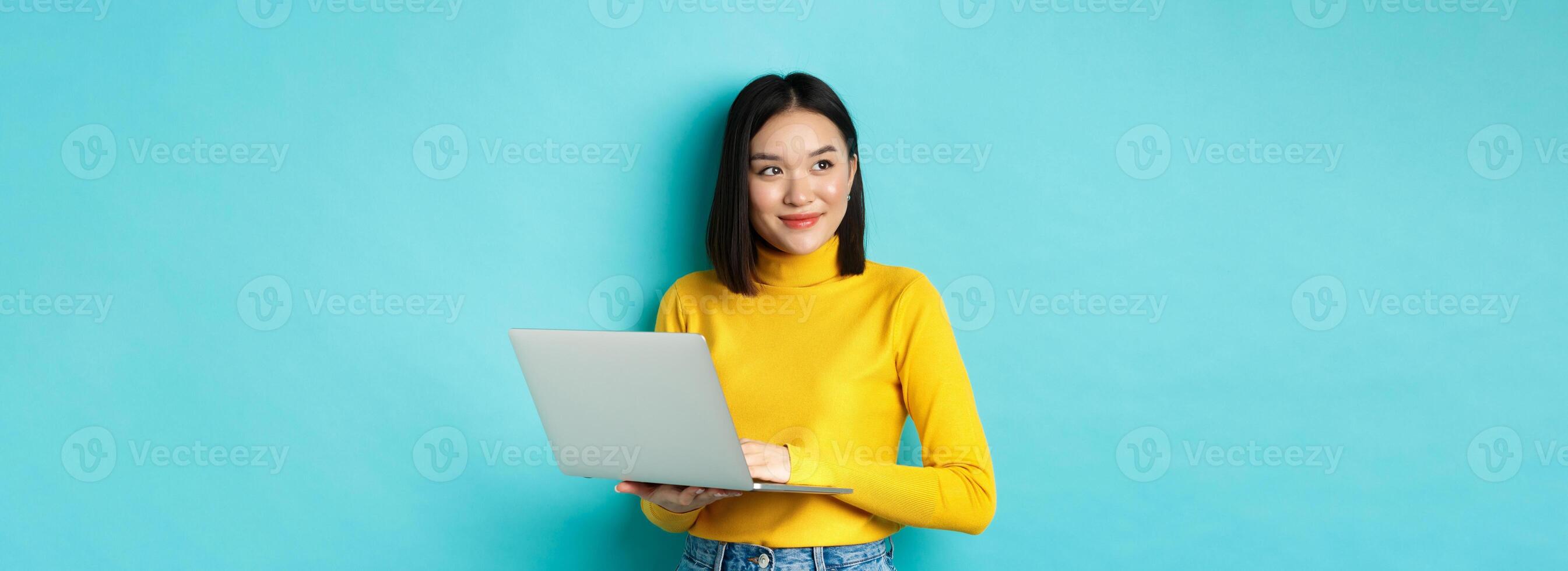 Image of stylish korean woman working on laptop and looking away with dreamy smile, standing with computer against blue background photo