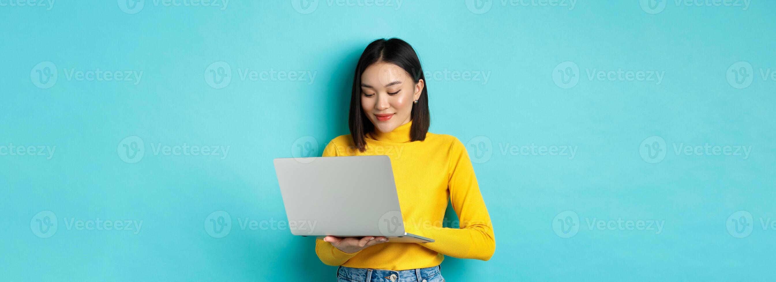 Beautiful asian female student working on laptop, typing on keyboard and looking at screen with pleased smile, standing over blue background photo