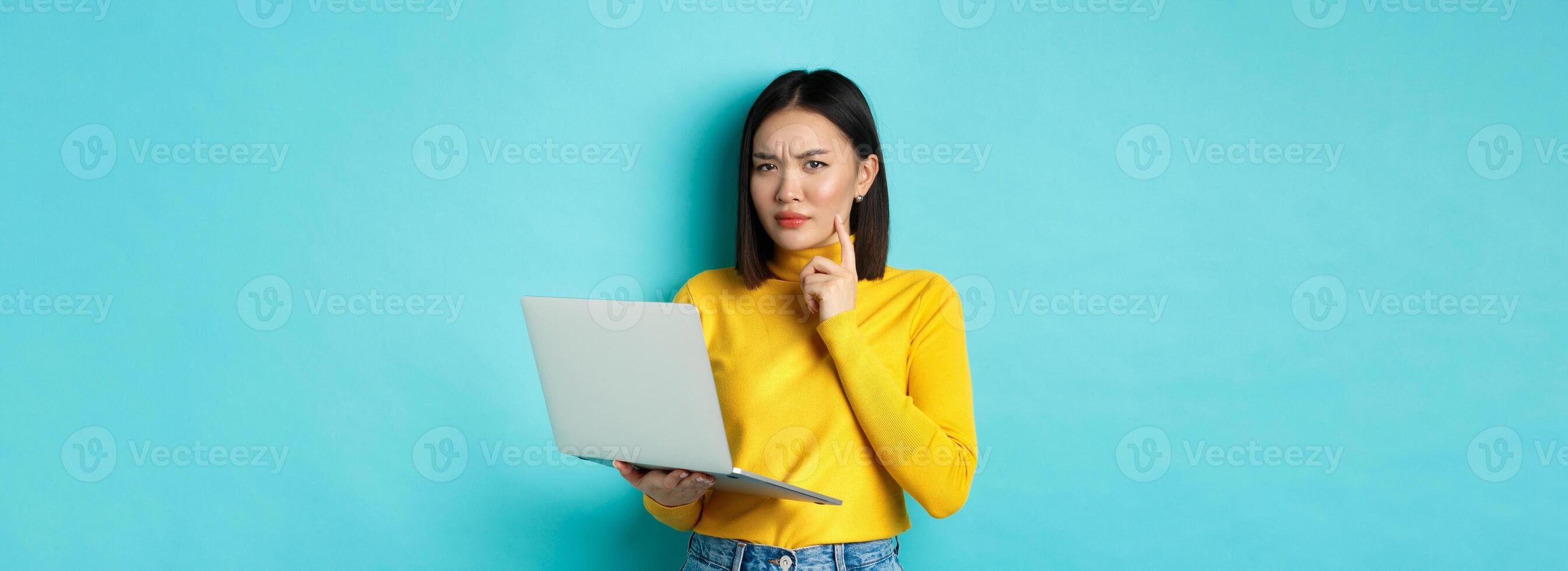 Serious looking asian woman working on laptop and thinking, frowning at camera, solving problem at work, standing over blue background photo