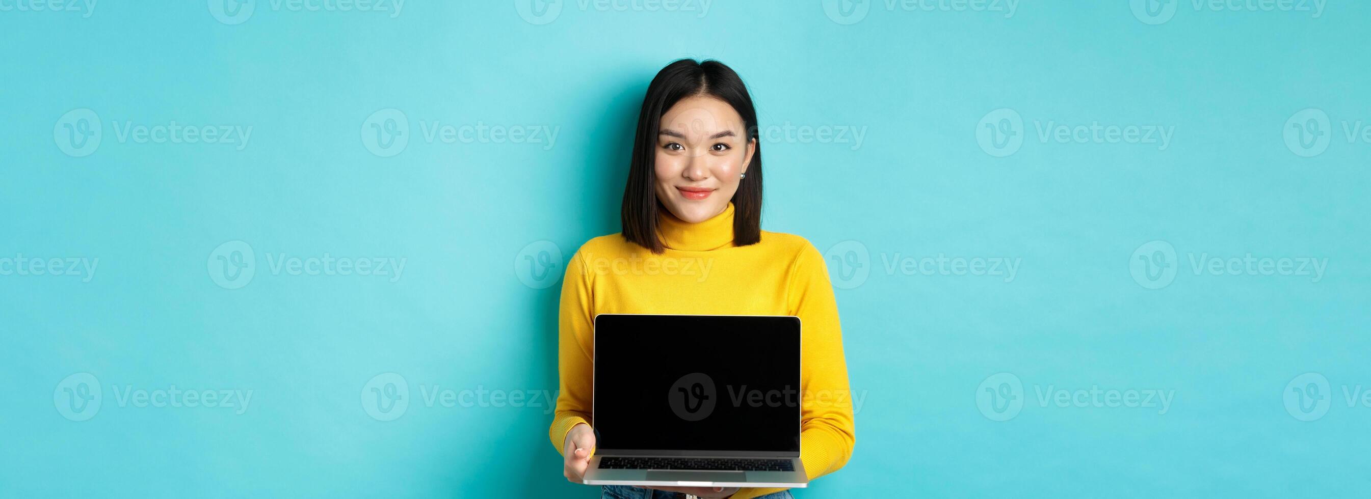 Beautiful and stylish asian woman demonstrate product on screen, showing empty laptop display and smiling, standing over blue background photo