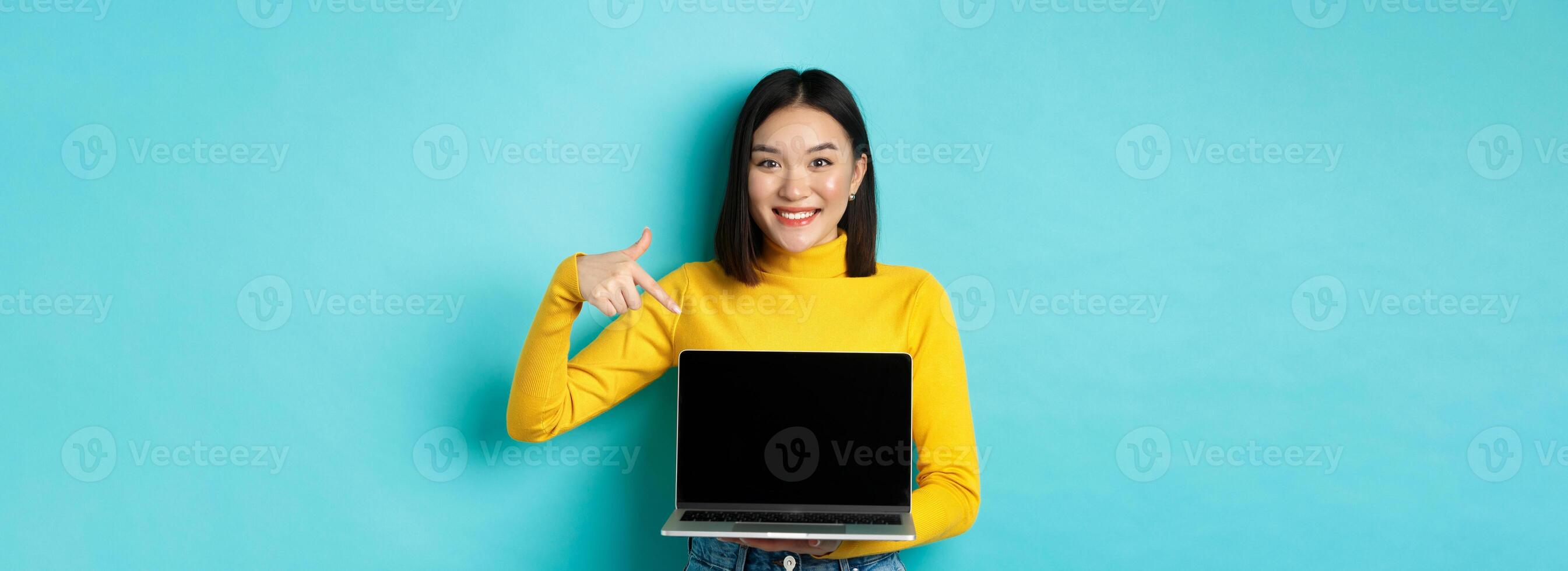 E-commerce and online shopping concept. Beautiful young woman pointing and showing laptop screen, smiling at camera, standing over blue background photo