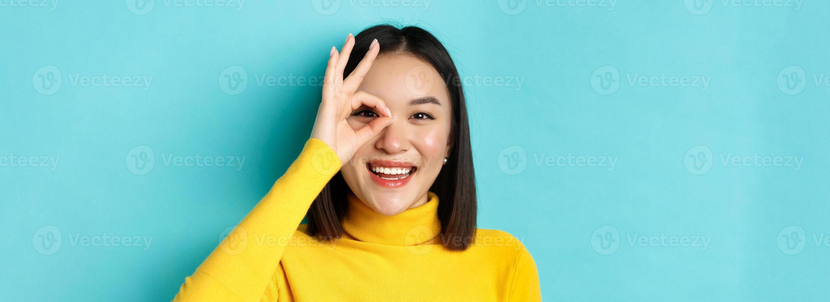 Beauty and makeup concept. Close up of carefree asian girl showing OK sign on eye and smiling, looking happy at camera, standing over blue background photo