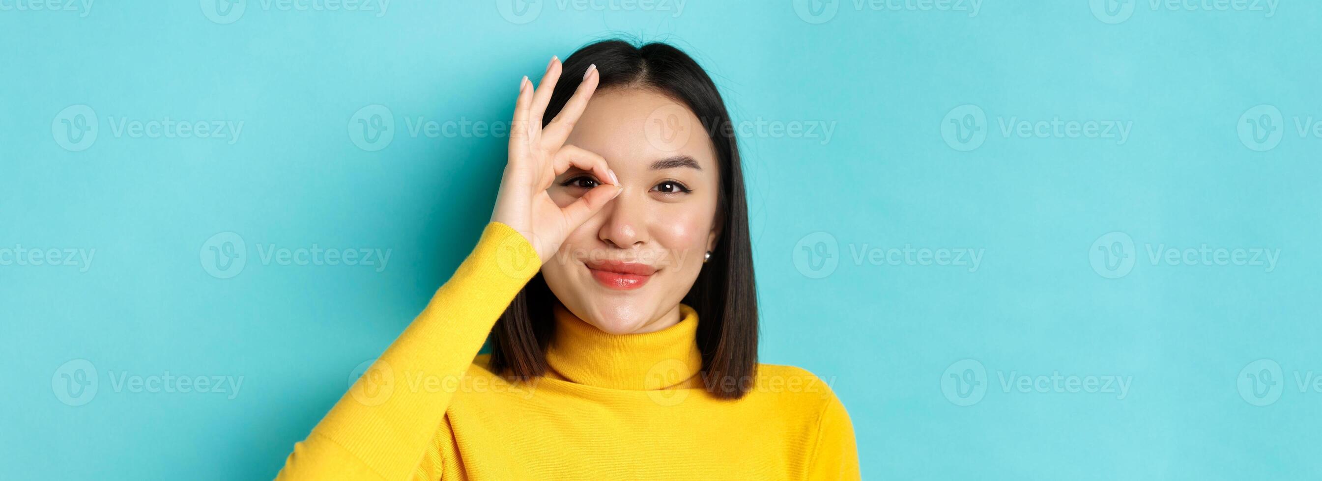 Beauty and makeup concept. Close up of carefree asian girl showing OK sign on eye and smiling, looking happy at camera, standing over blue background photo