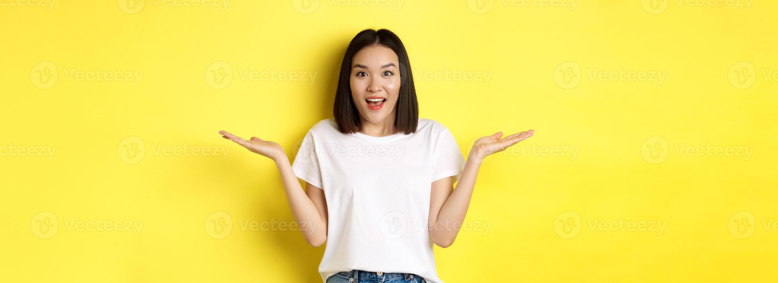 Beauty and fashion concept. Surprised asian girl spread hands sideways, holding on palms, pointing at two products, standing over yellow background photo