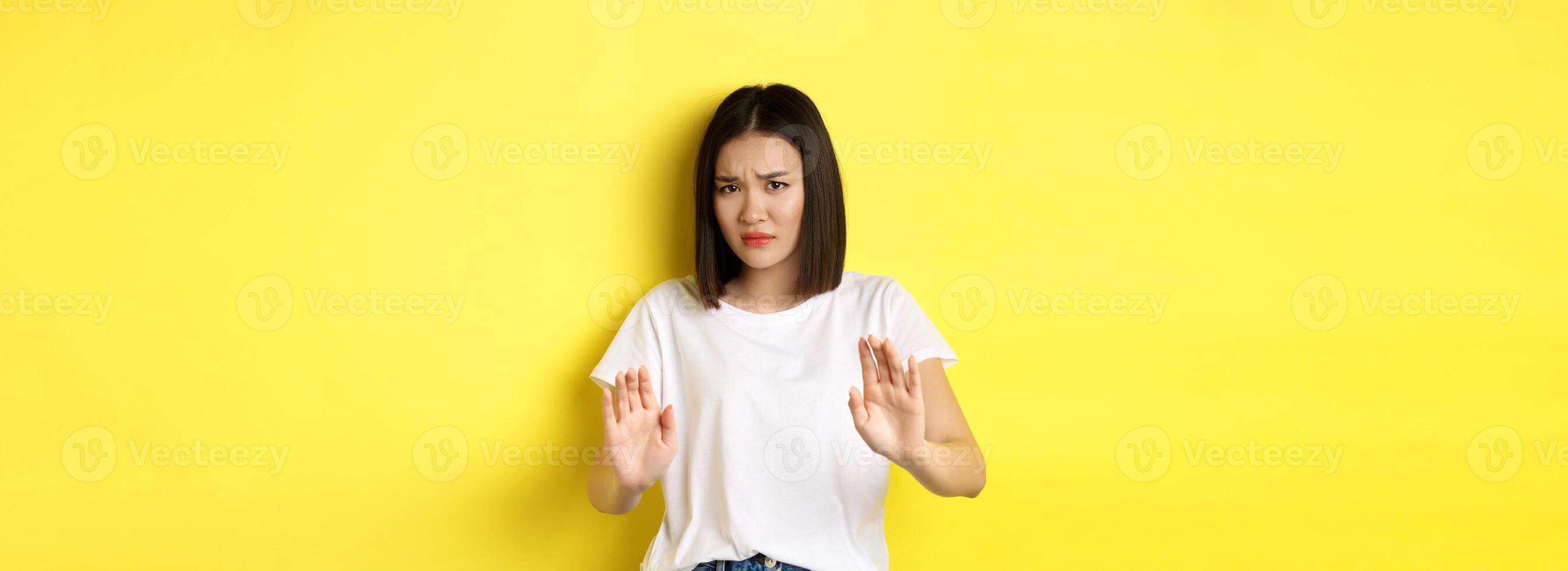 Please no. Asian woman, victim of assualt or domestic abuse, pleading with hands raised up in defense, frowning sad, begging stop, standing over yellow background photo
