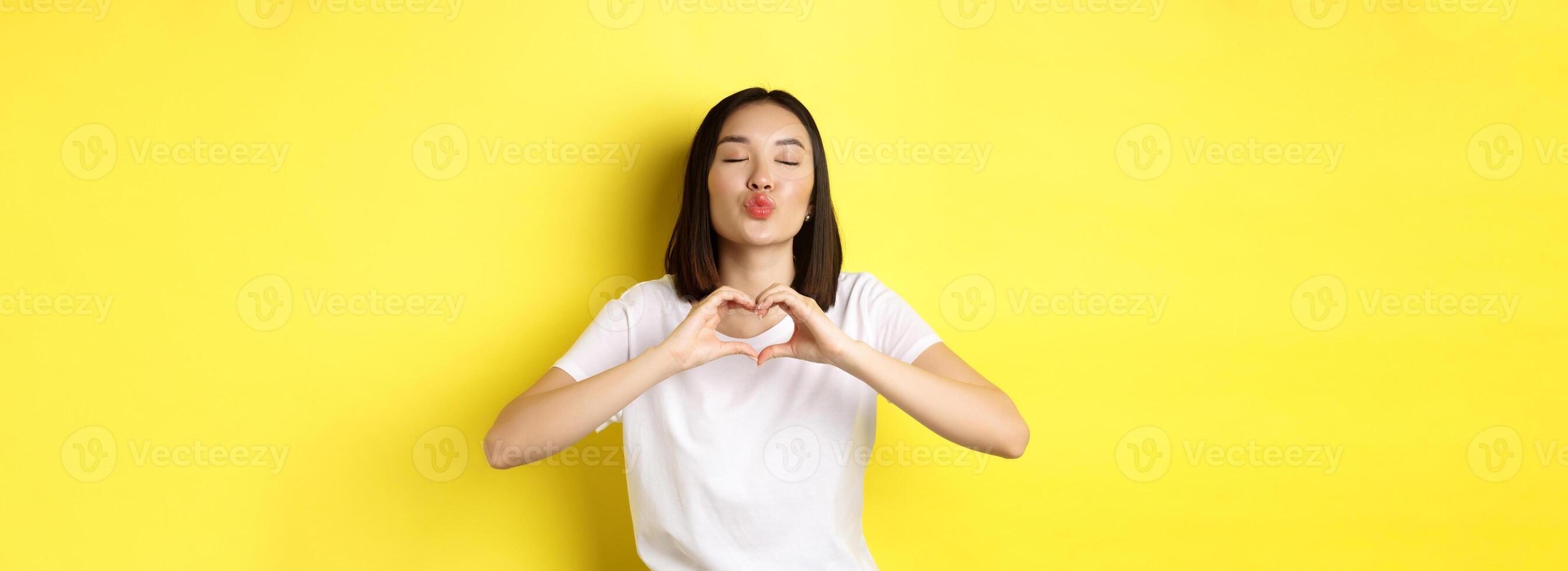 Beautiful asian woman showing I love you heart gesture, smiling at camera, standing against yellow background. Concept of valentines day and romance photo