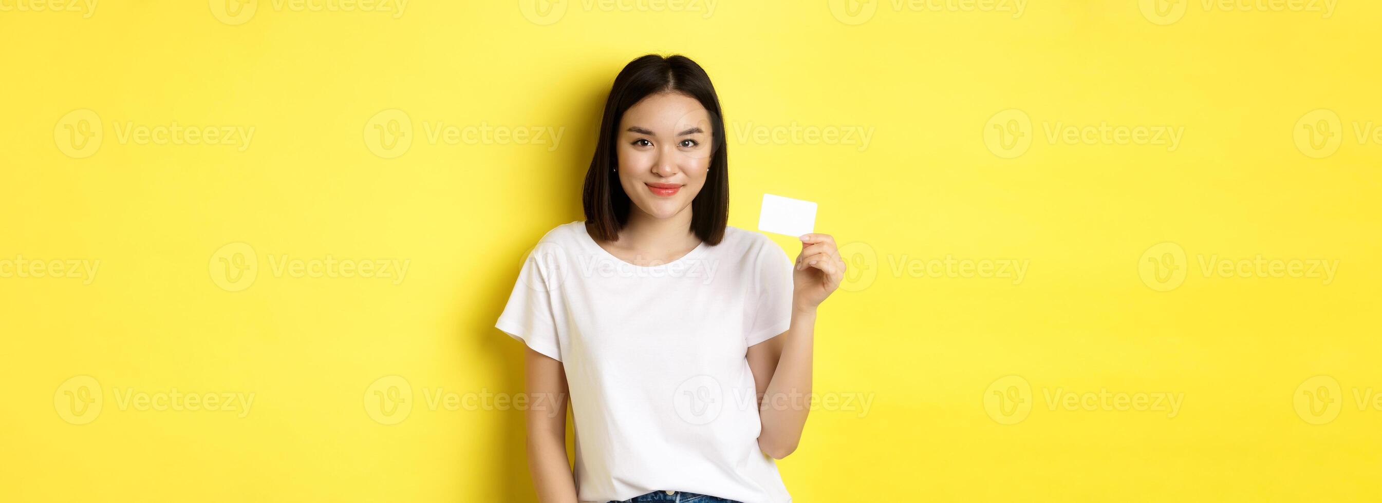 Young asian woman in casual white t-shirt showing plastic credit card and smiling at camera, yellow background photo