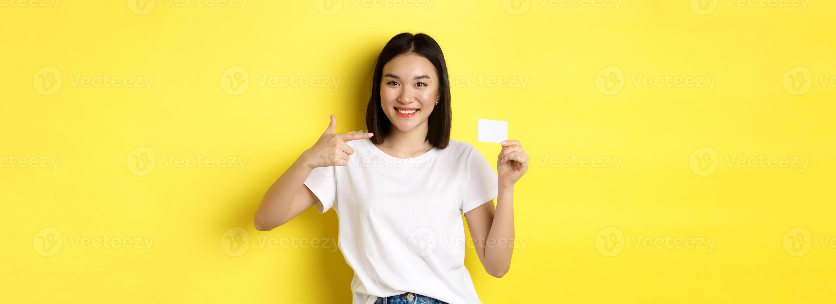 Young asian woman in casual white t-shirt showing plastic credit card and smiling at camera, yellow background photo